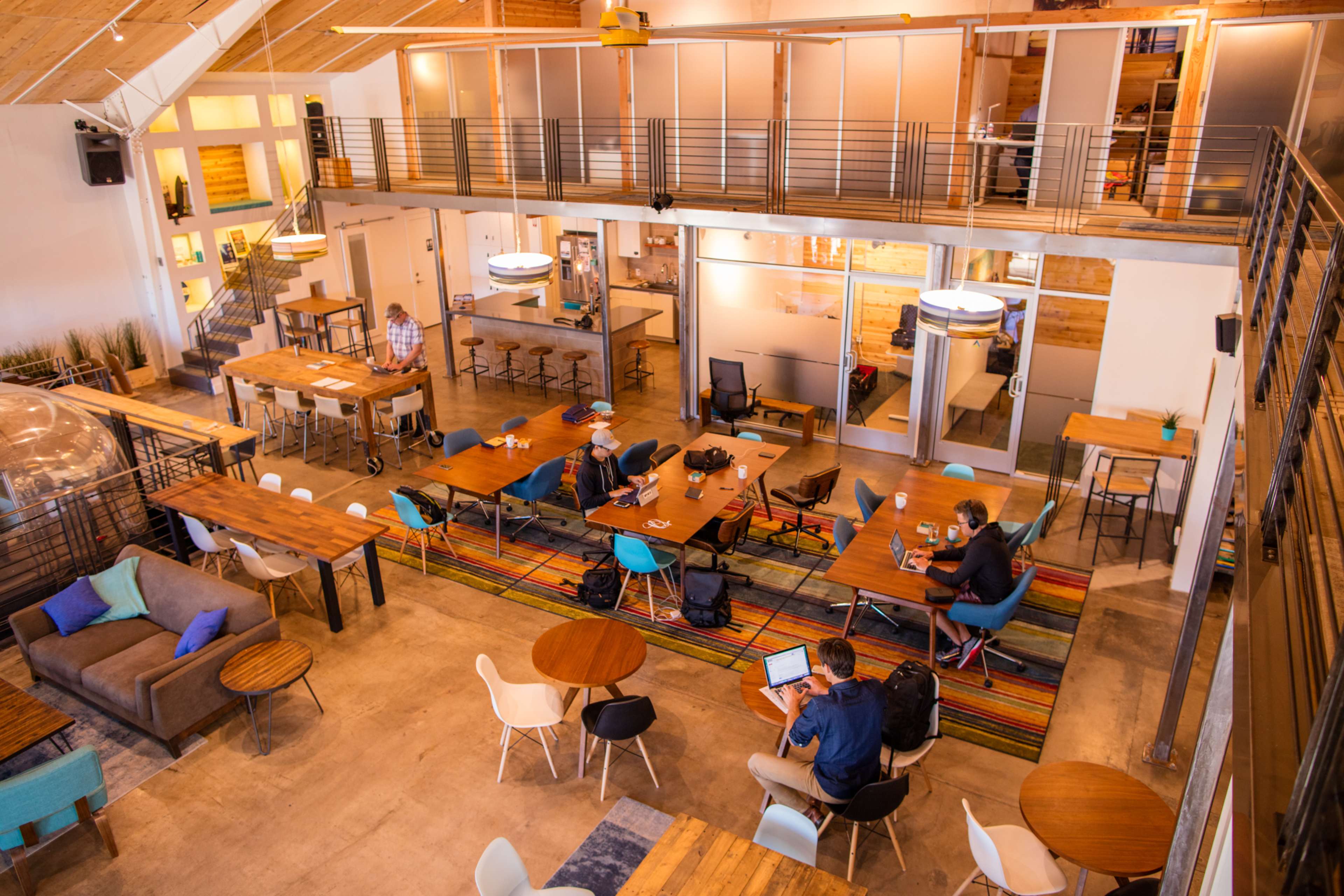 A spacious co-working space features multiple tables, a lounge area, and individuals working on laptops.