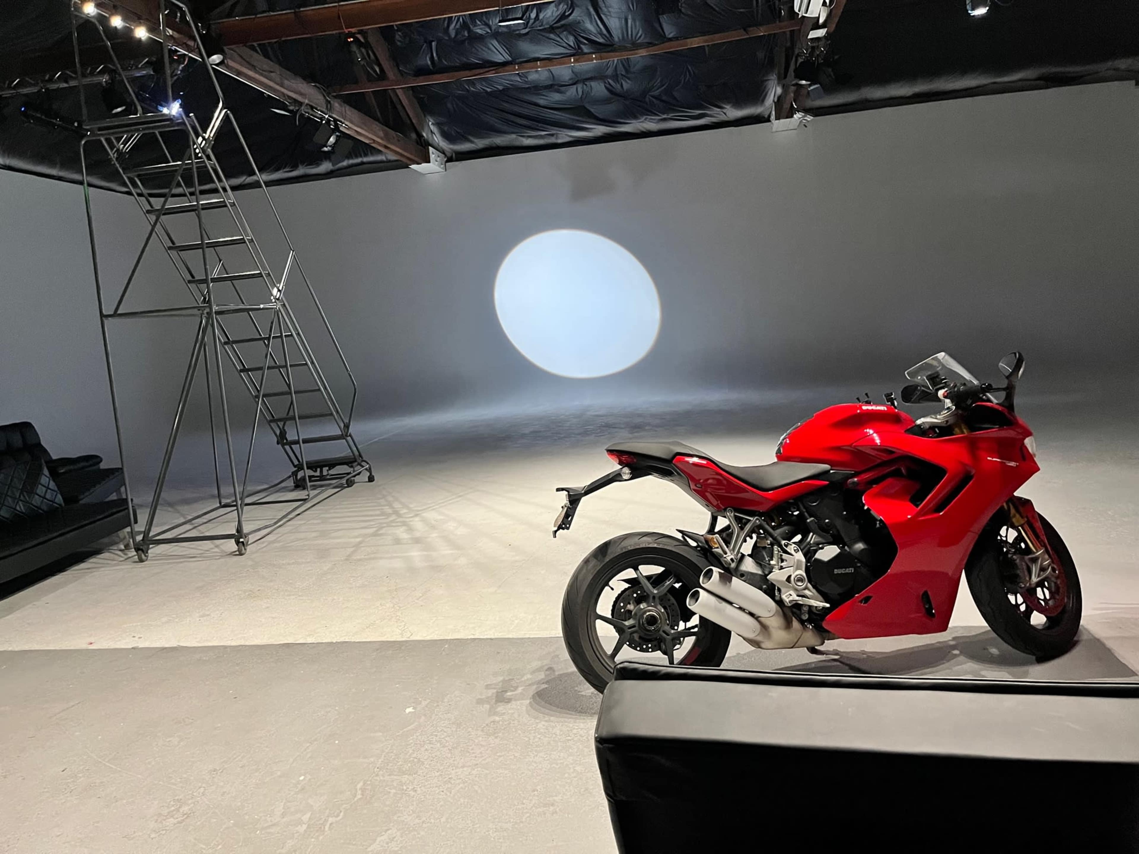 A red motorcycle is positioned beside a black couch in a spacious studio with a circular light projection on the floor.