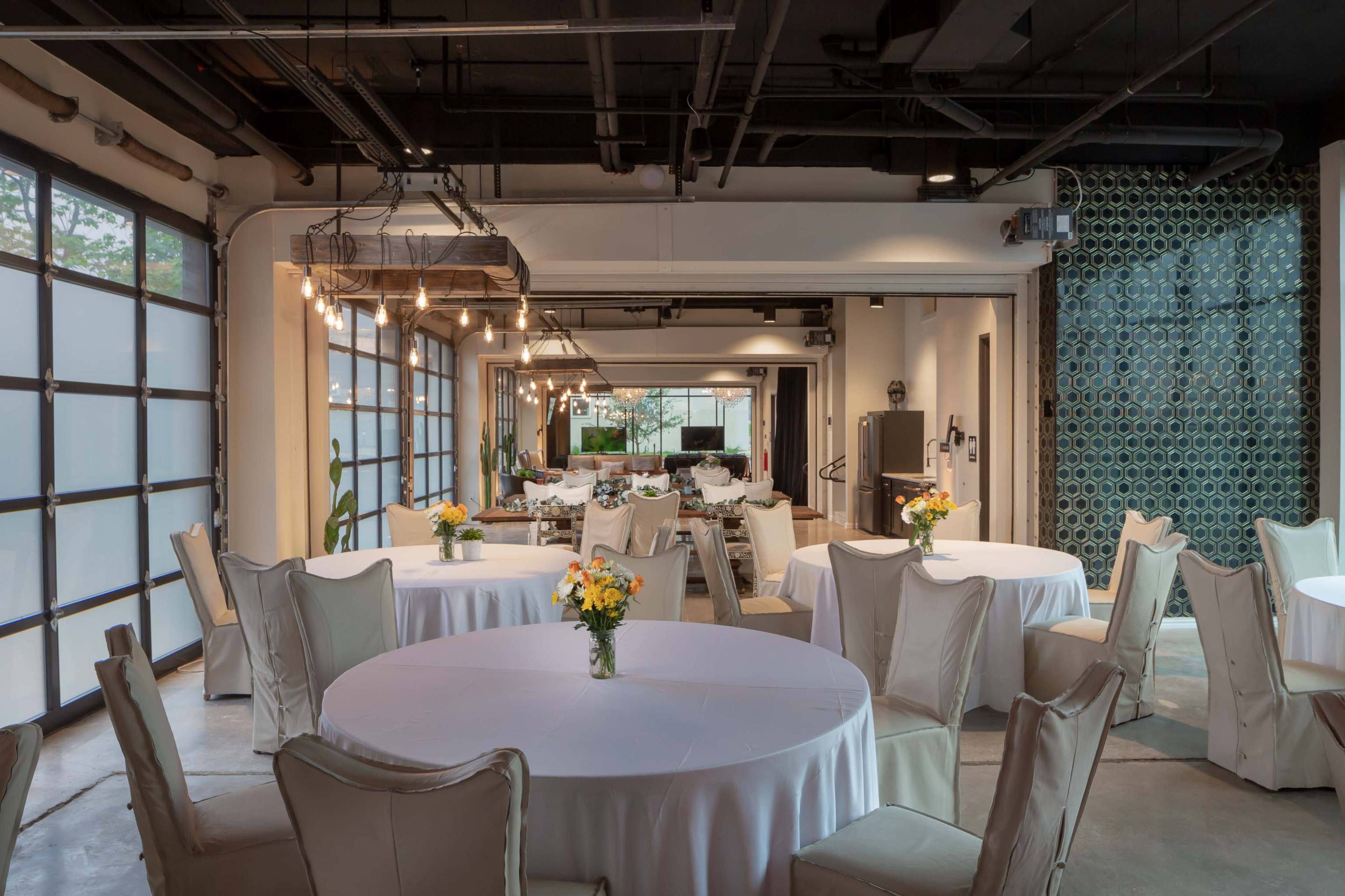 A dining area with round tables covered in white tablecloths, decorated with small floral centerpieces, and large windows allowing natural light to enter.