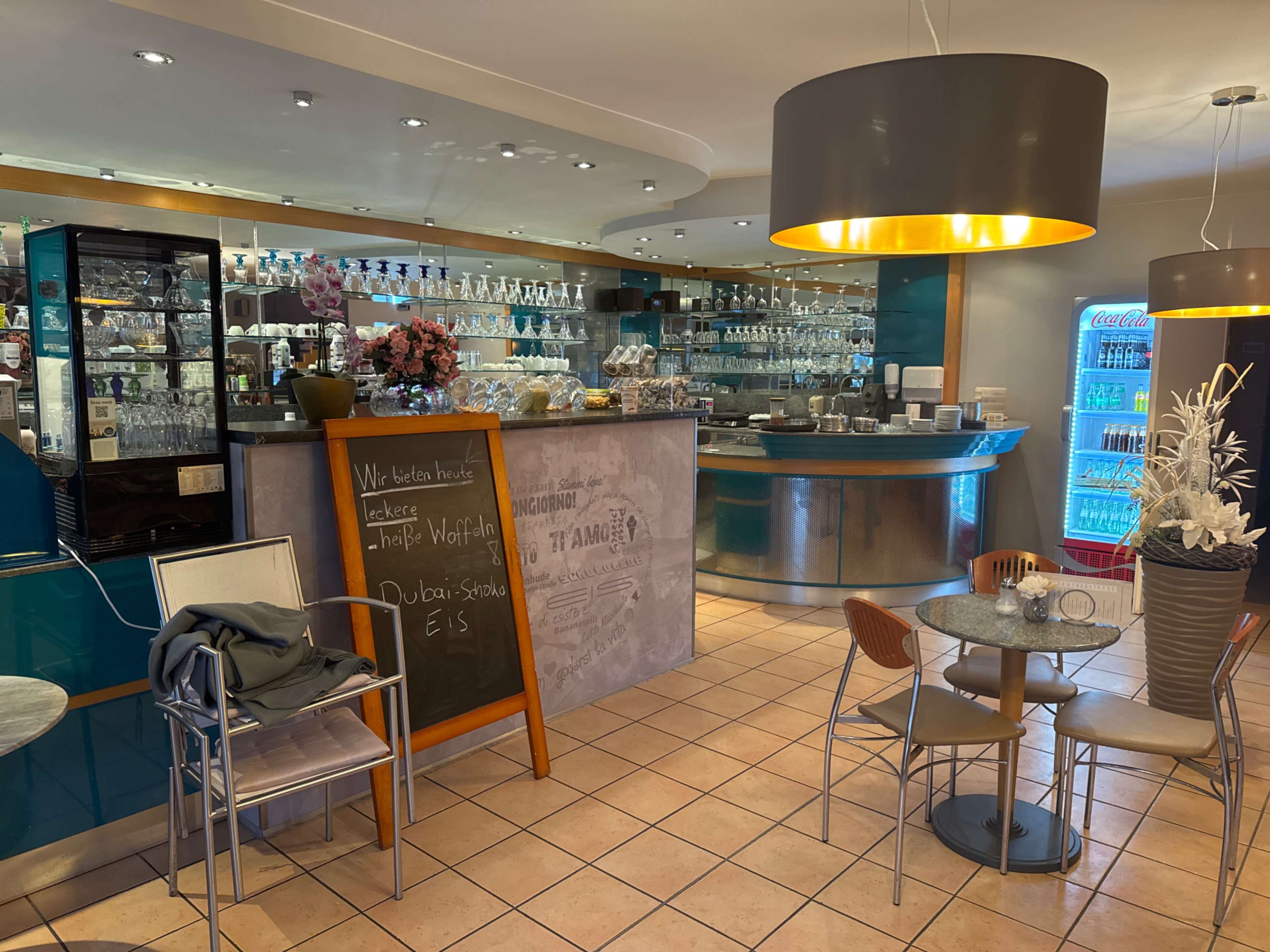 The image shows a modern café interior with a stylish bar counter, glass shelving displaying glassware, and a seating area featuring round tables and chairs.