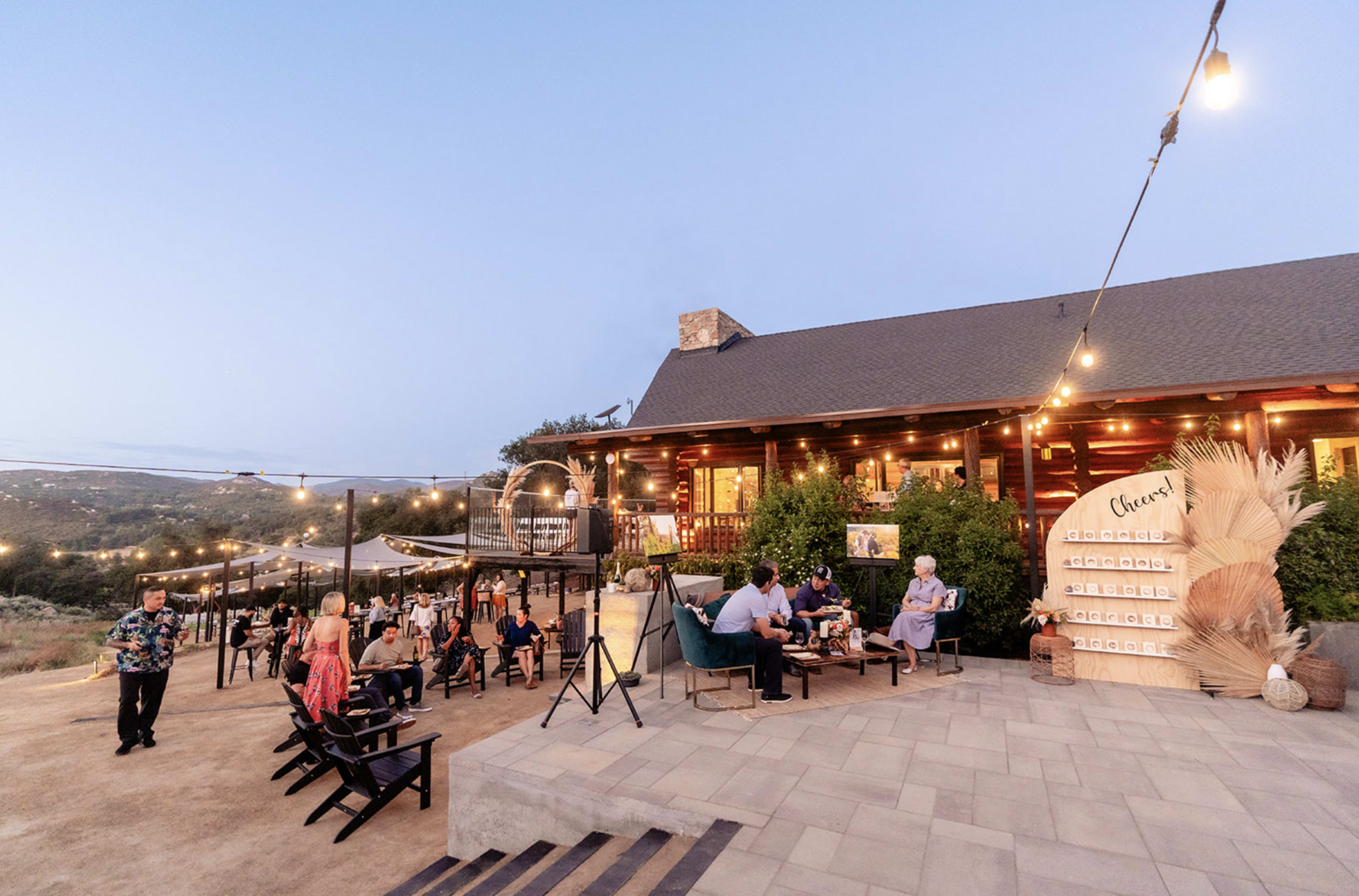 A gathering of people takes place outside a rustic-style building, adorned with string lights and surrounded by greenery, as they engage in conversation and activities on a stone patio.