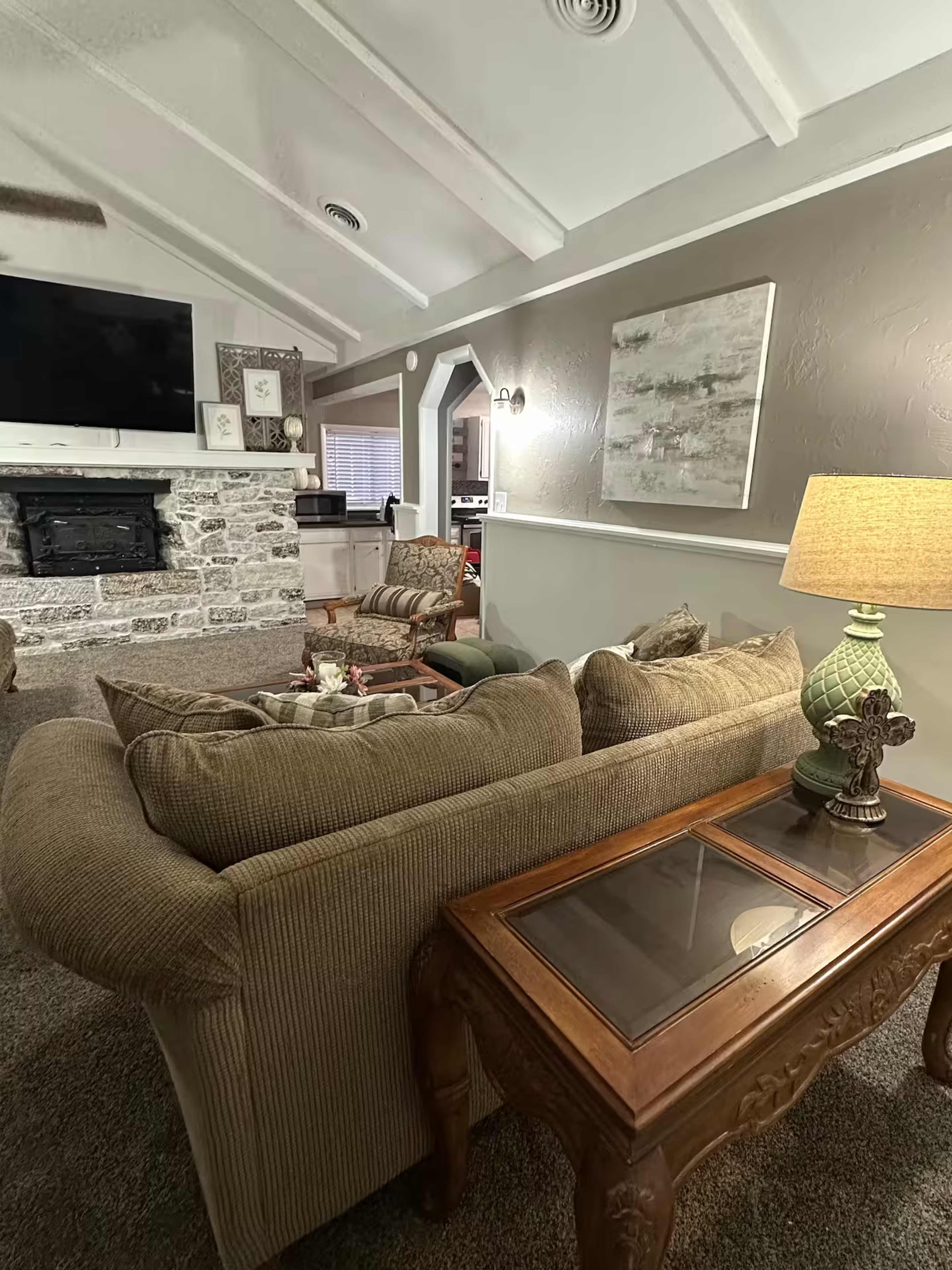 The image shows a cozy living room with a large sectional sofa, a wooden coffee table, and a stone fireplace, all illuminated by a table lamp.