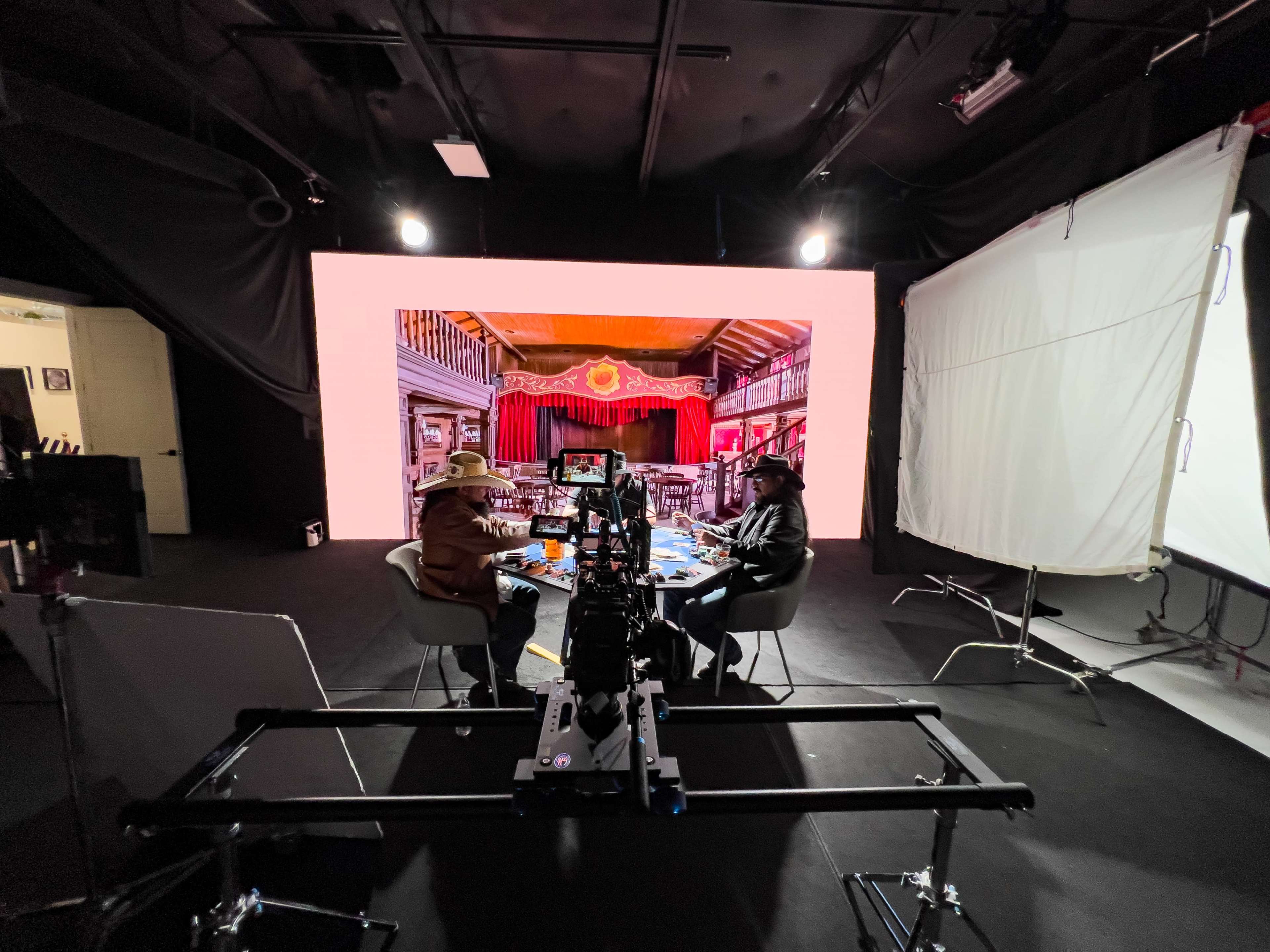 A film crew is set up in a black studio with a large backdrop displaying an ornate interior scene while two individuals sit at a table in front of the camera.