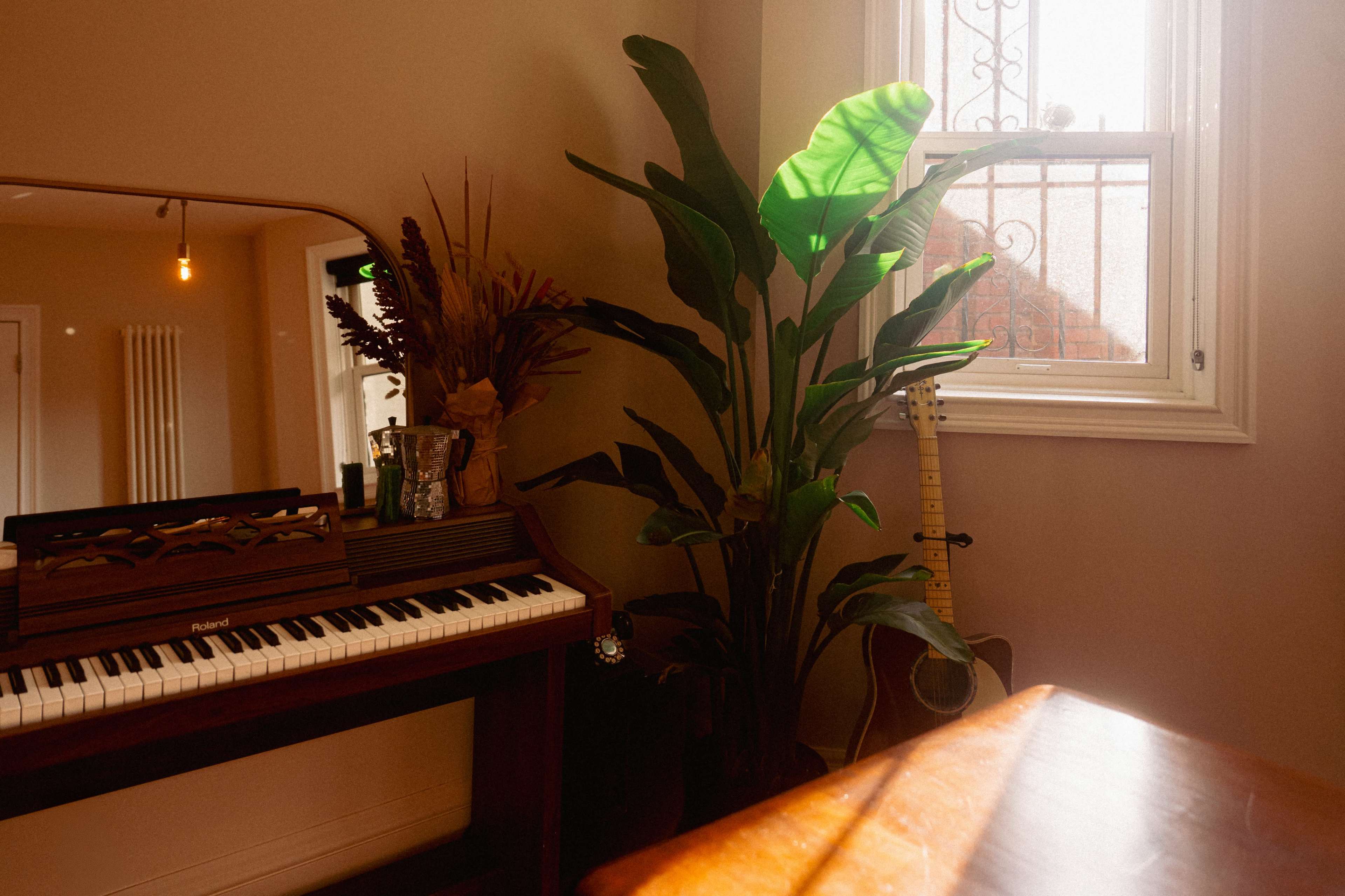 A wooden piano with a mirror above it sits next to a tall potted plant and an acoustic guitar against the wall, illuminated by natural light from a nearby window.