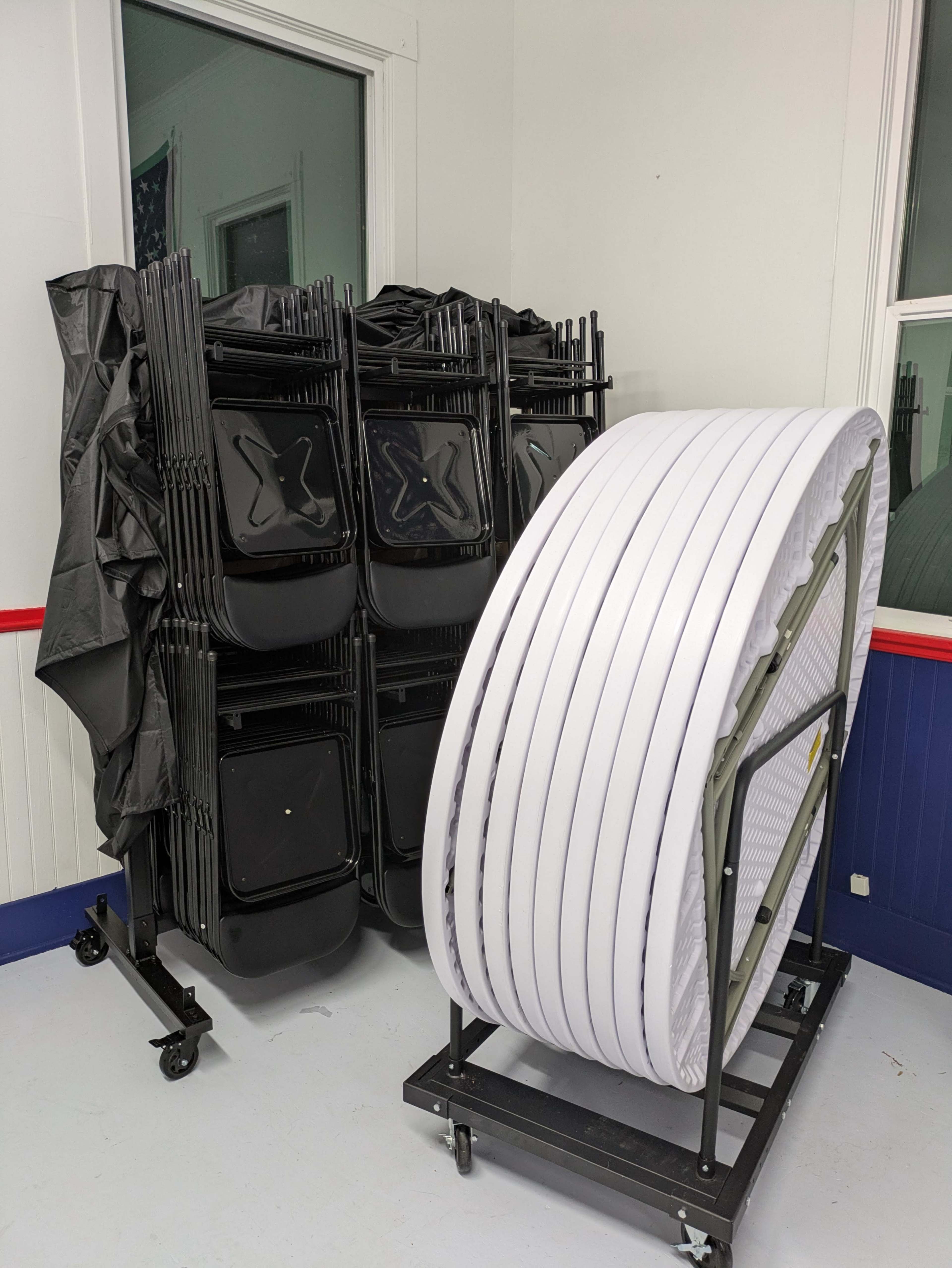 The image shows a stack of black folding chairs beside a cart containing rolled white tables in a room with white walls and blue accents.