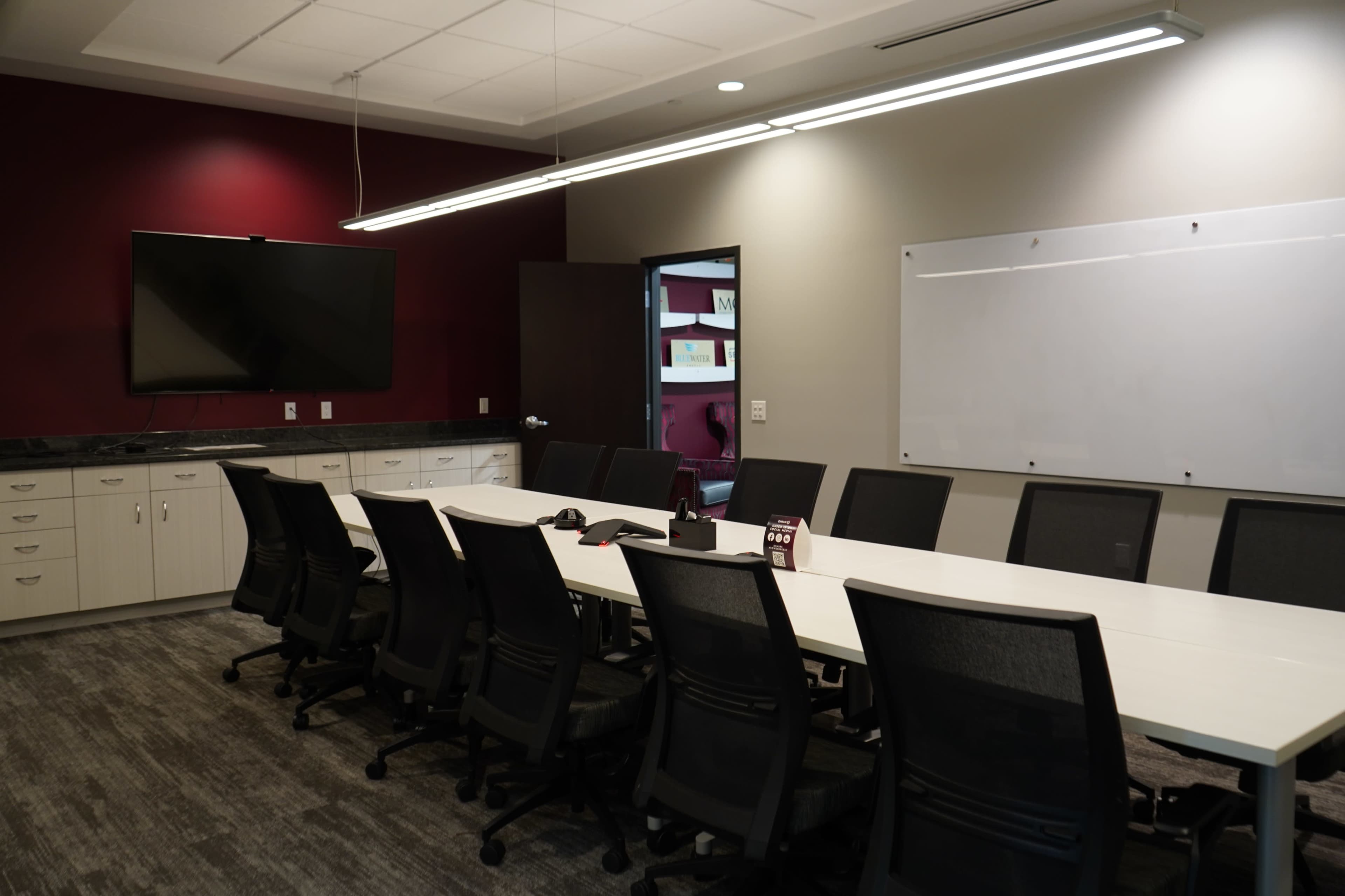 A modern conference room features a long table surrounded by black ergonomic chairs, a large wall-mounted television, and a whiteboard on one side.