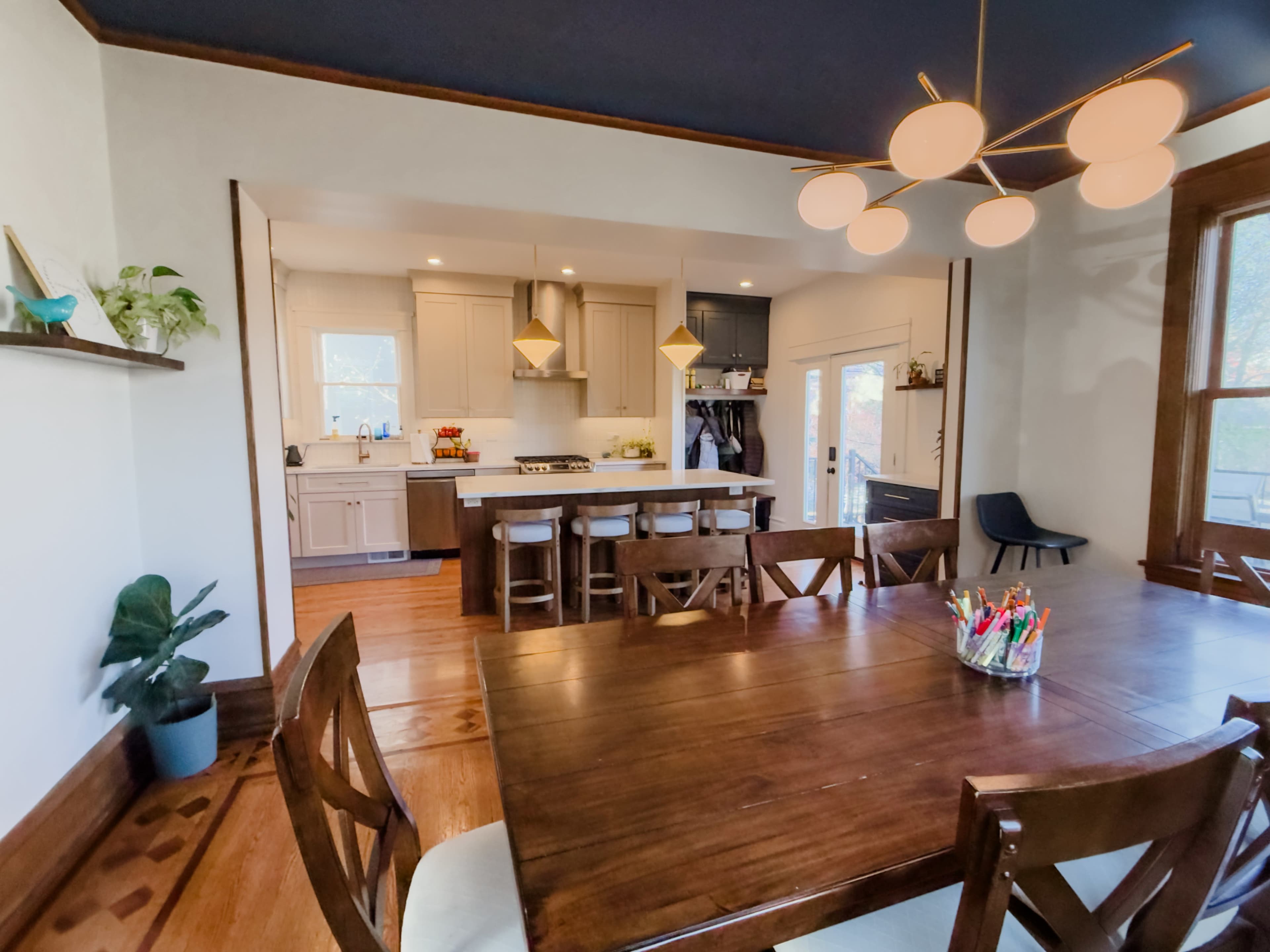 A dining room features a large wooden table with chairs and an open view into a modern kitchen with light cabinetry and a large window.