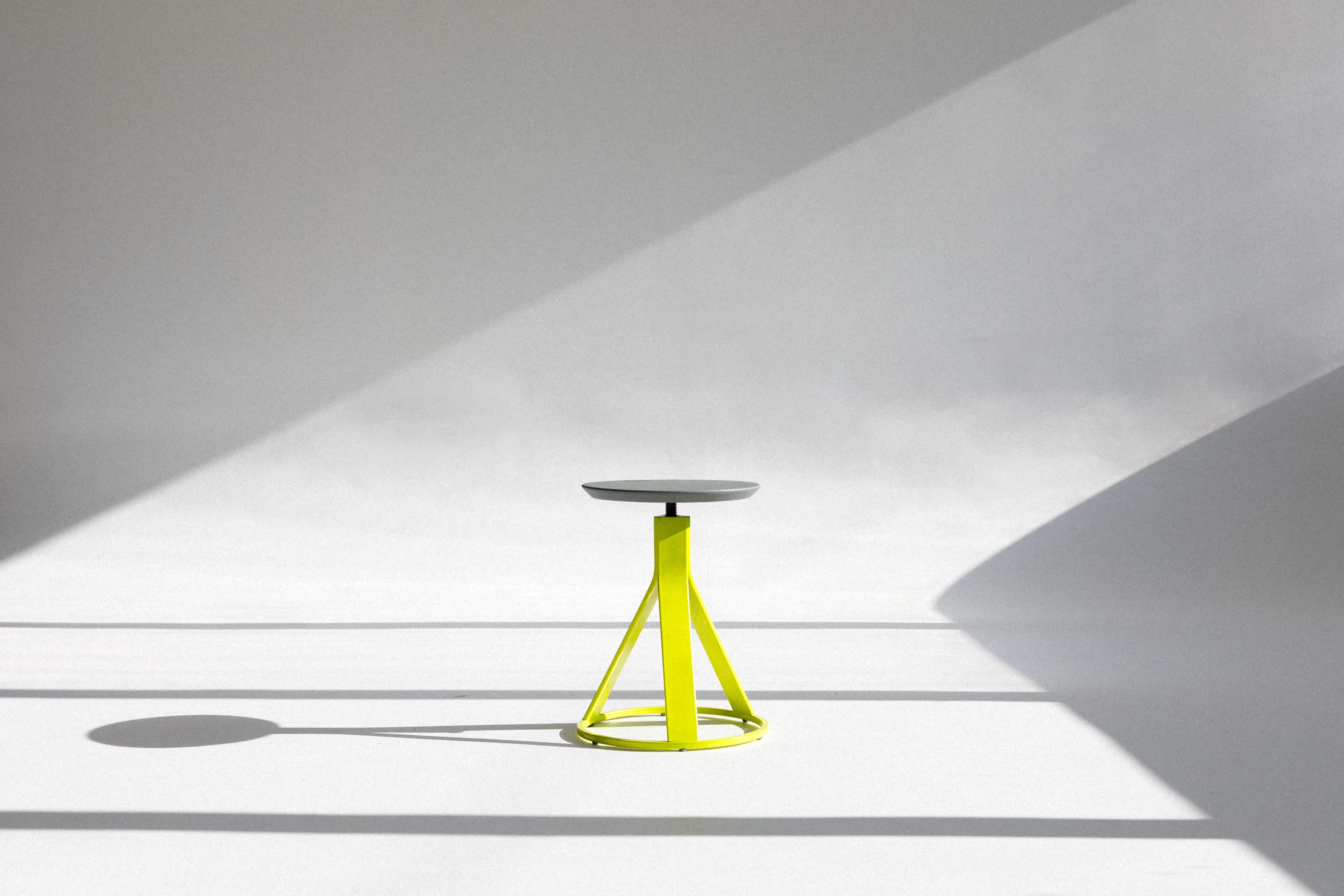 A simple stool with a gray seat and bright yellow legs sits on a white floor beneath soft, diffused light.