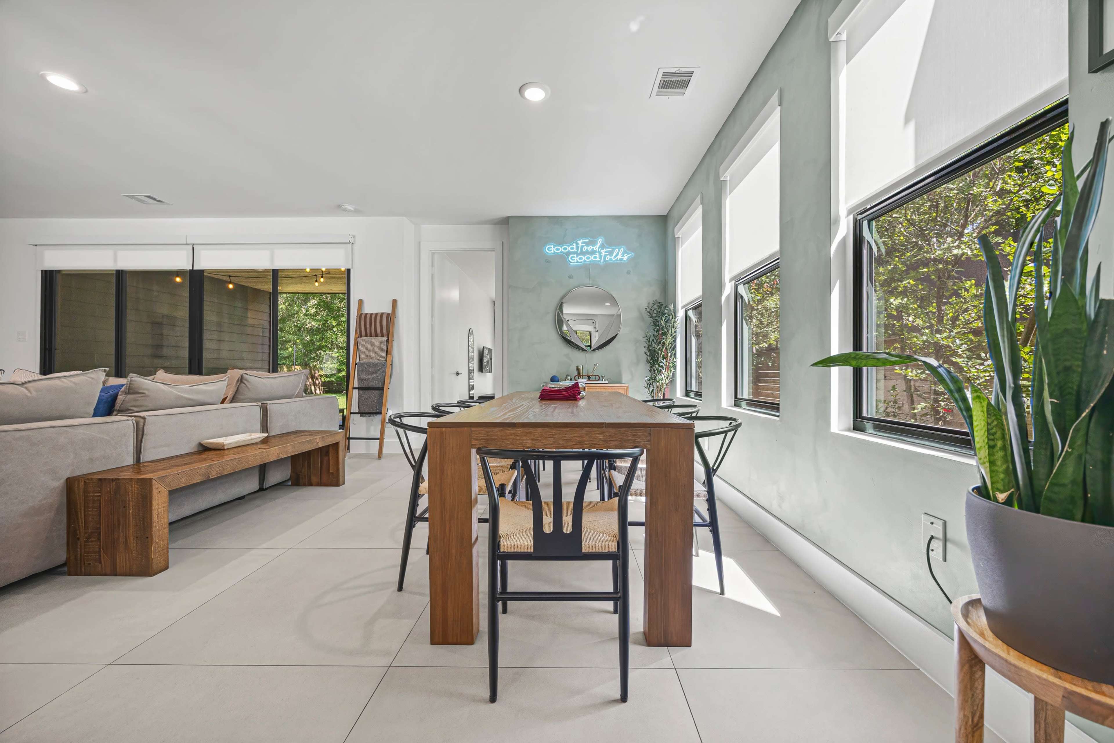 A dining area features a wooden table surrounded by black chairs, positioned near large windows that allow natural light to fill the space, while a couch is visible in the background.