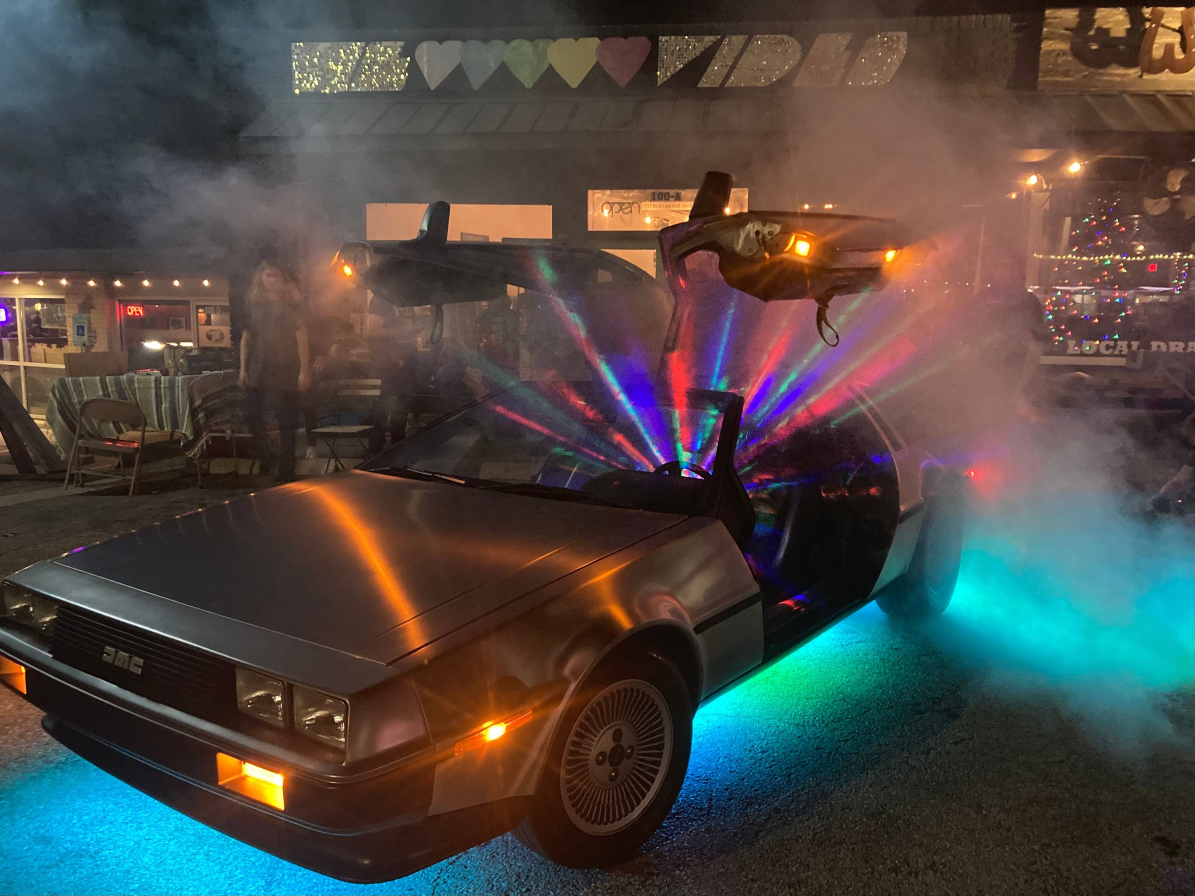 A DeLorean car with its doors open sits amidst colorful lights and fog at a nighttime event.