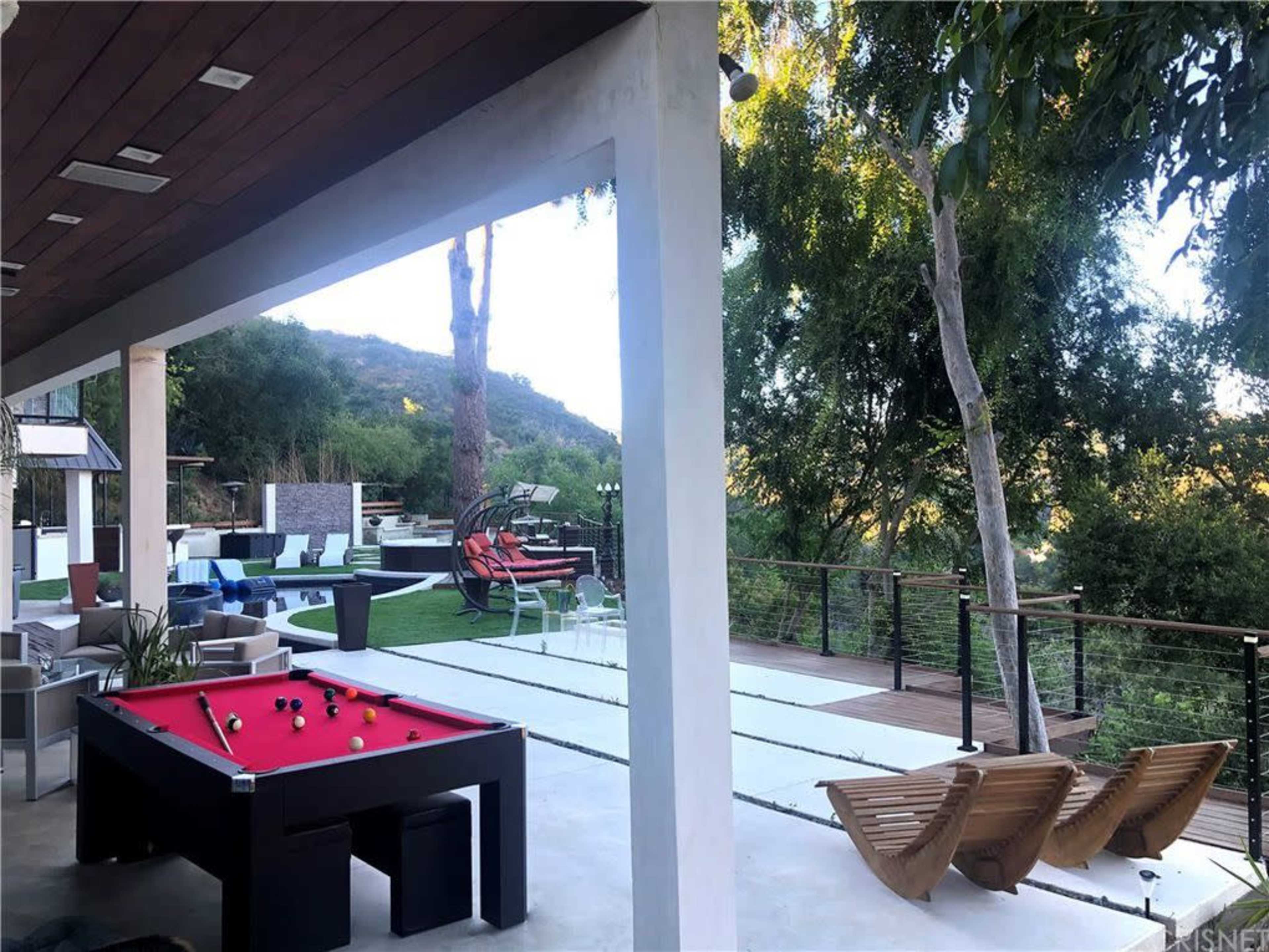 A modern outdoor space featuring a red pool table, wooden lounge chairs, and a landscaped area with greenery in the background.