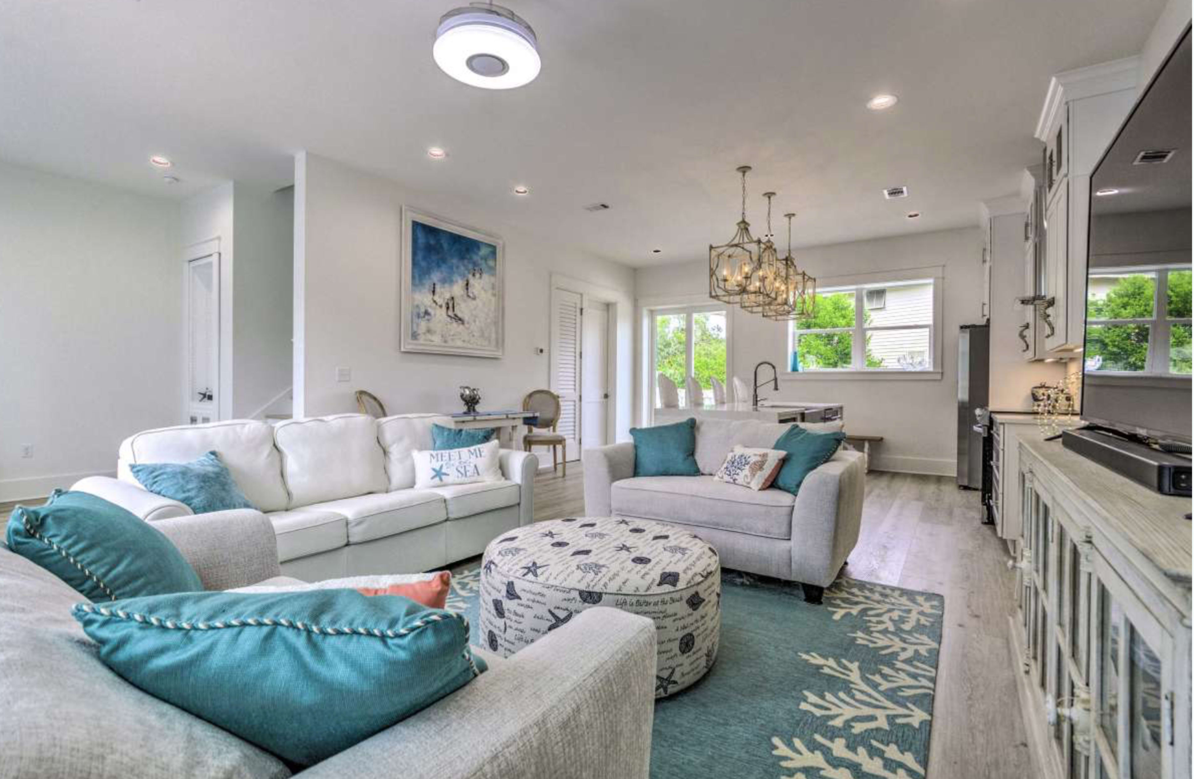 A bright and modern living room featuring a white sofa, a patterned area rug, a circular ottoman, and a stylish dining area with a chandelier.