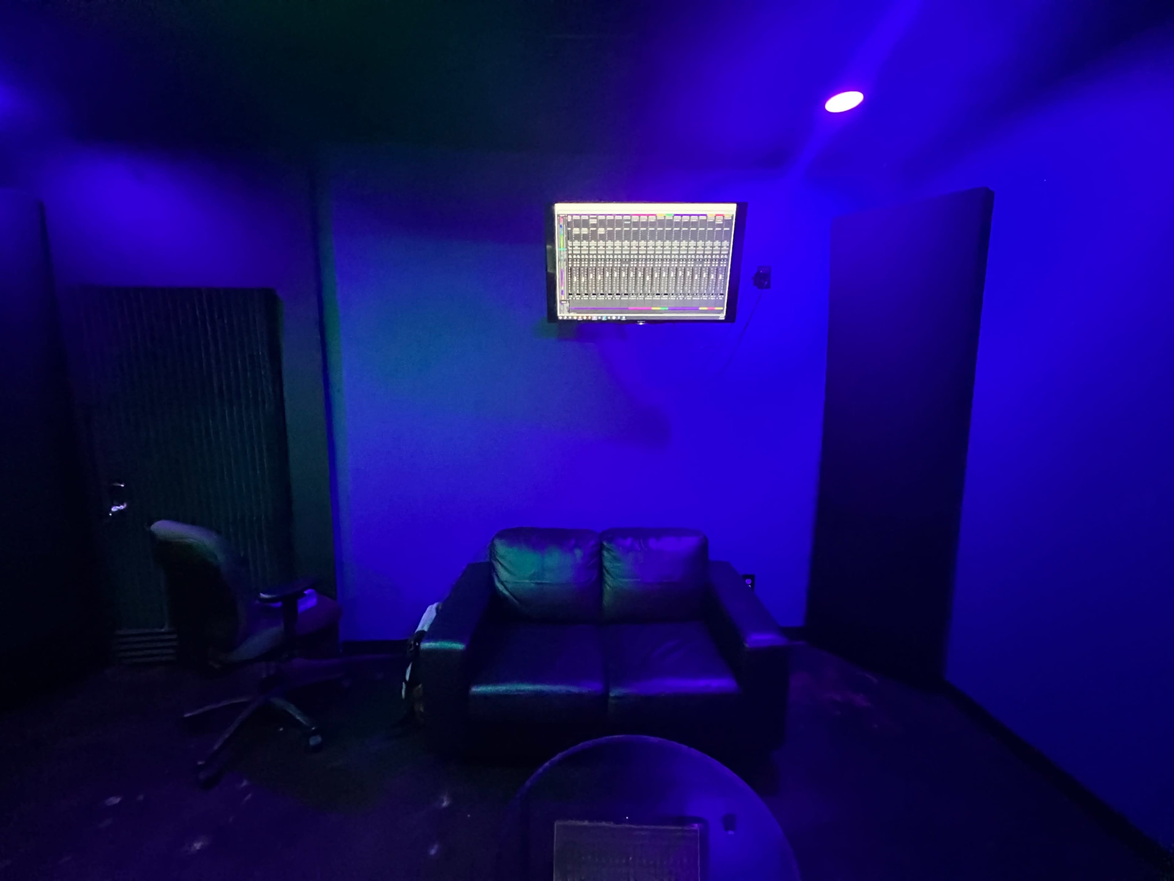 A dimly lit room with purple walls, a couch, a chair, and a monitor displaying sound mixing software mounted on the wall.