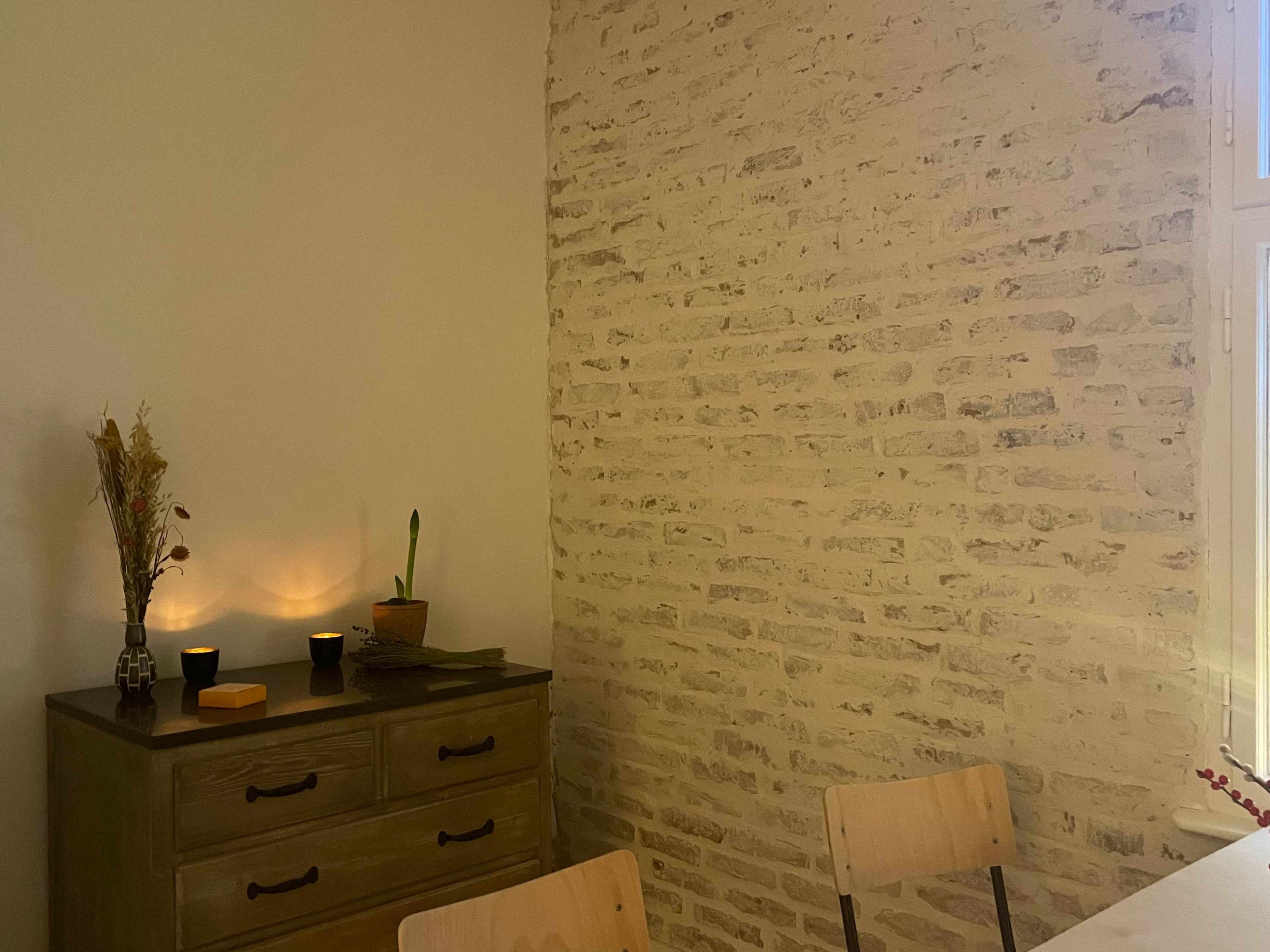 A minimalist interior with a wooden dresser, small plant, and candles casting a soft glow against a light-colored brick wall.