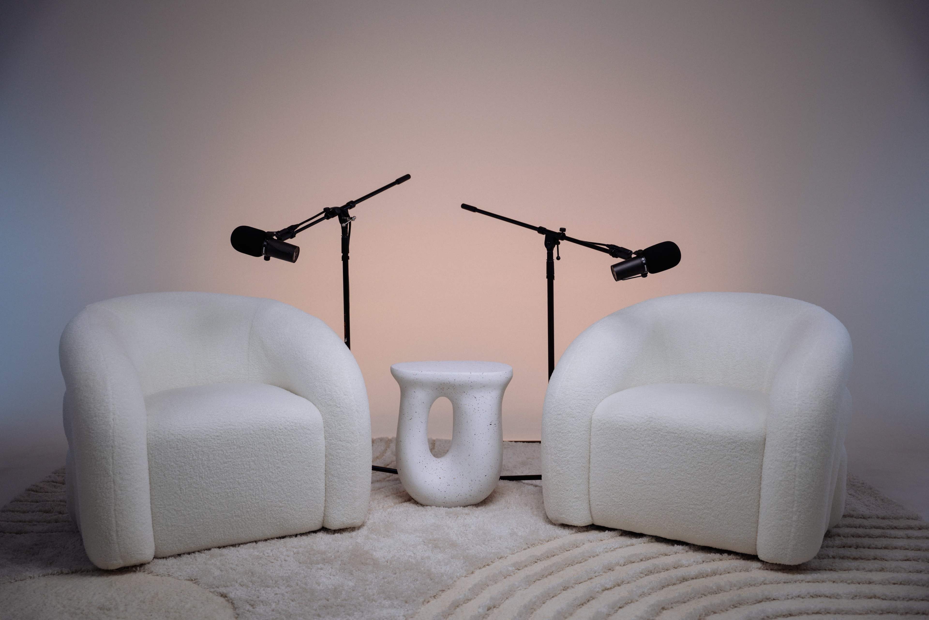 Two white, plush chairs are positioned facing each other with a small, round table in between, and two microphones mounted on stands nearby.