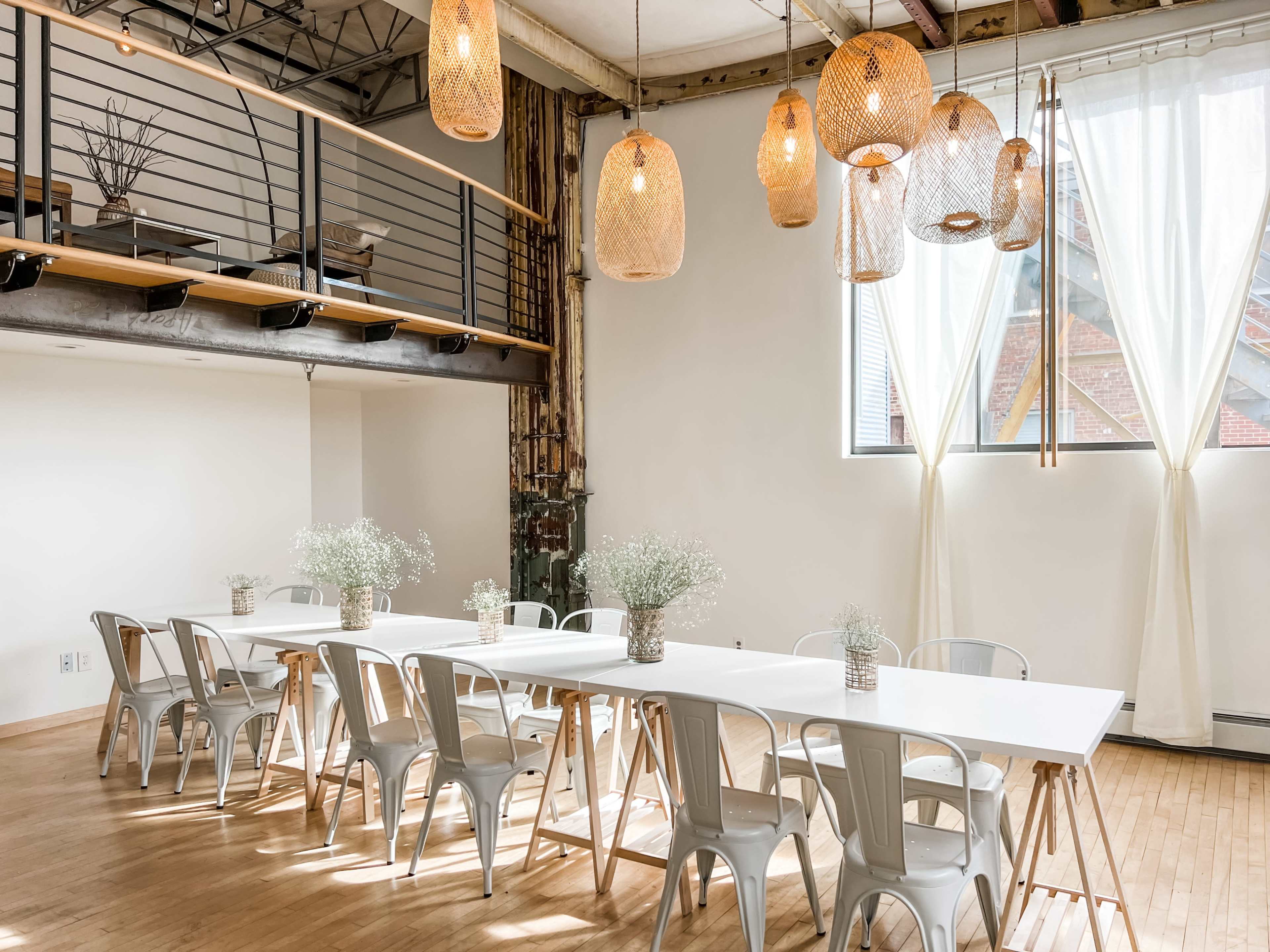 A long, white table with metal chairs is set up in a bright room featuring pendant lights and large windows, with simple floral arrangements on the table.