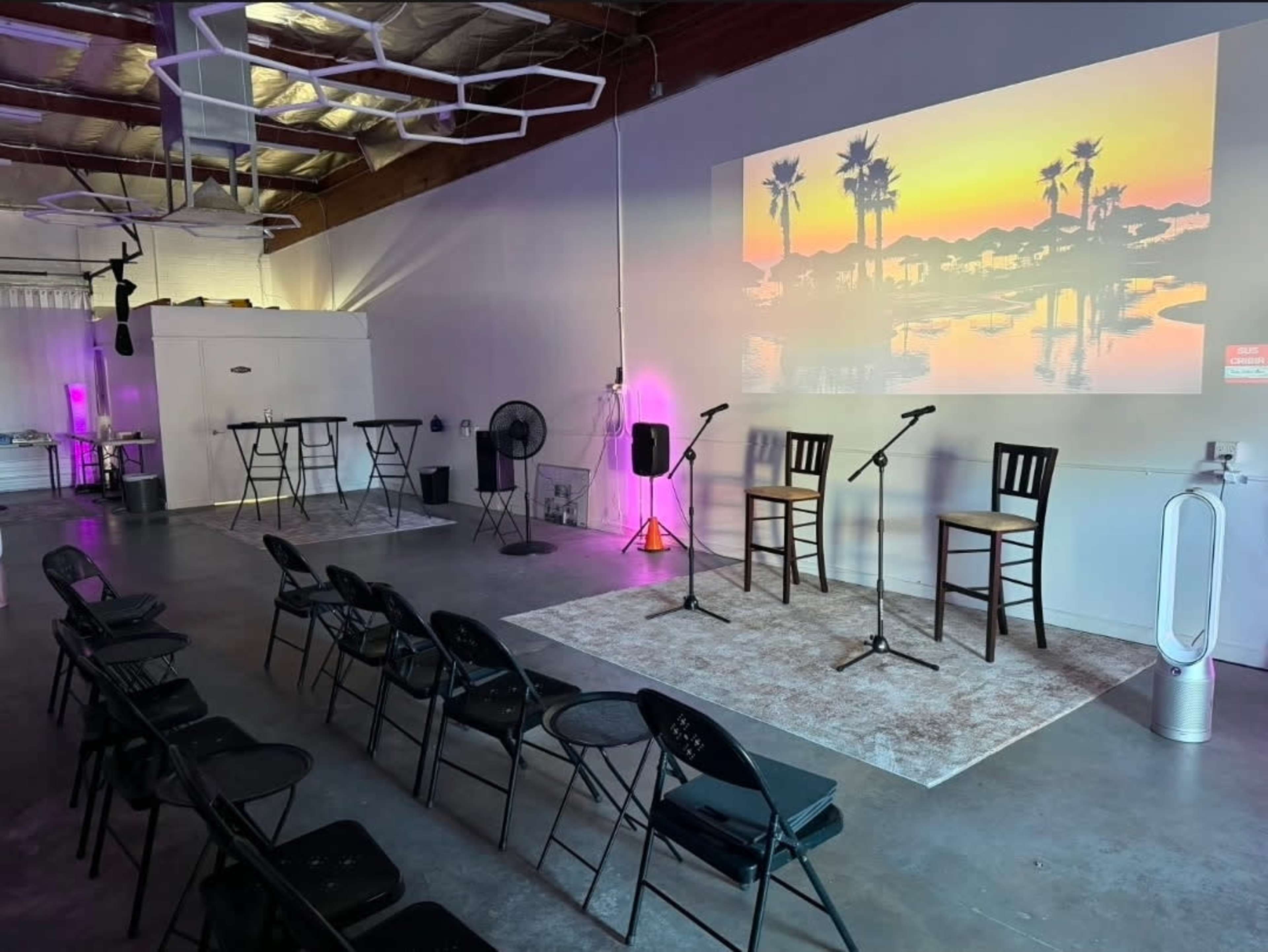 The image shows a spacious indoor area set up for an event, featuring two high chairs, microphones, and a large projected image of a sunset over palm trees on the wall.