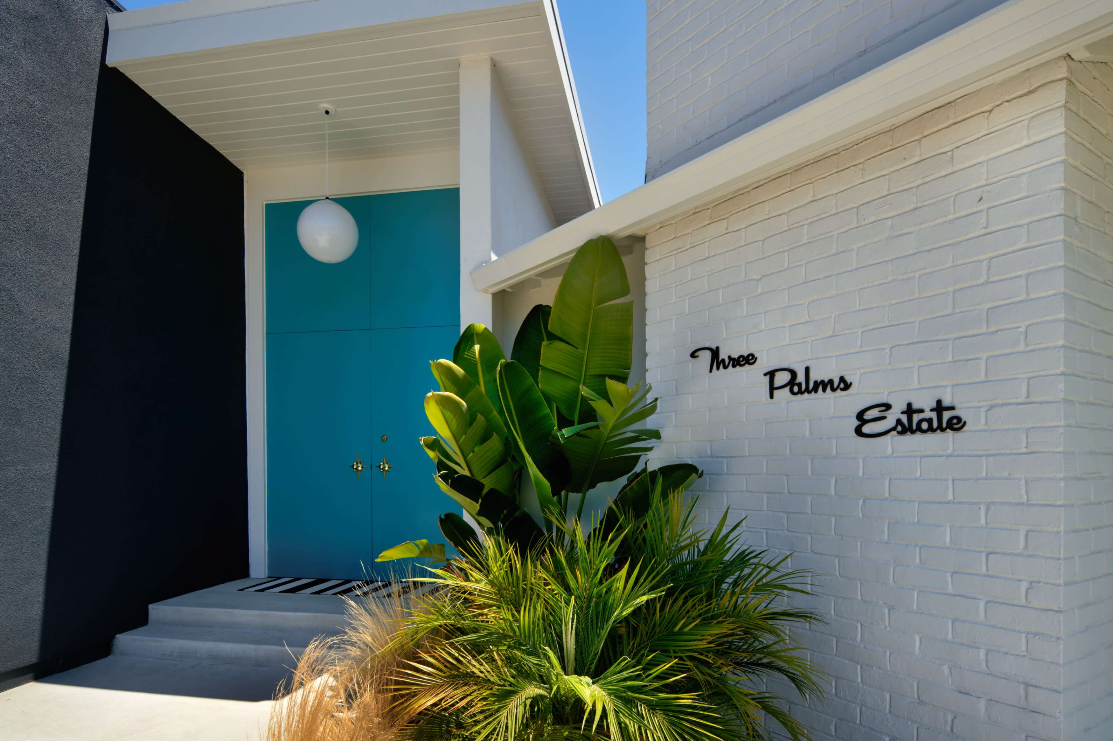 A modern entryway features a vibrant teal door next to a white brick wall with the words "Three Palms Estate" and lush greenery.