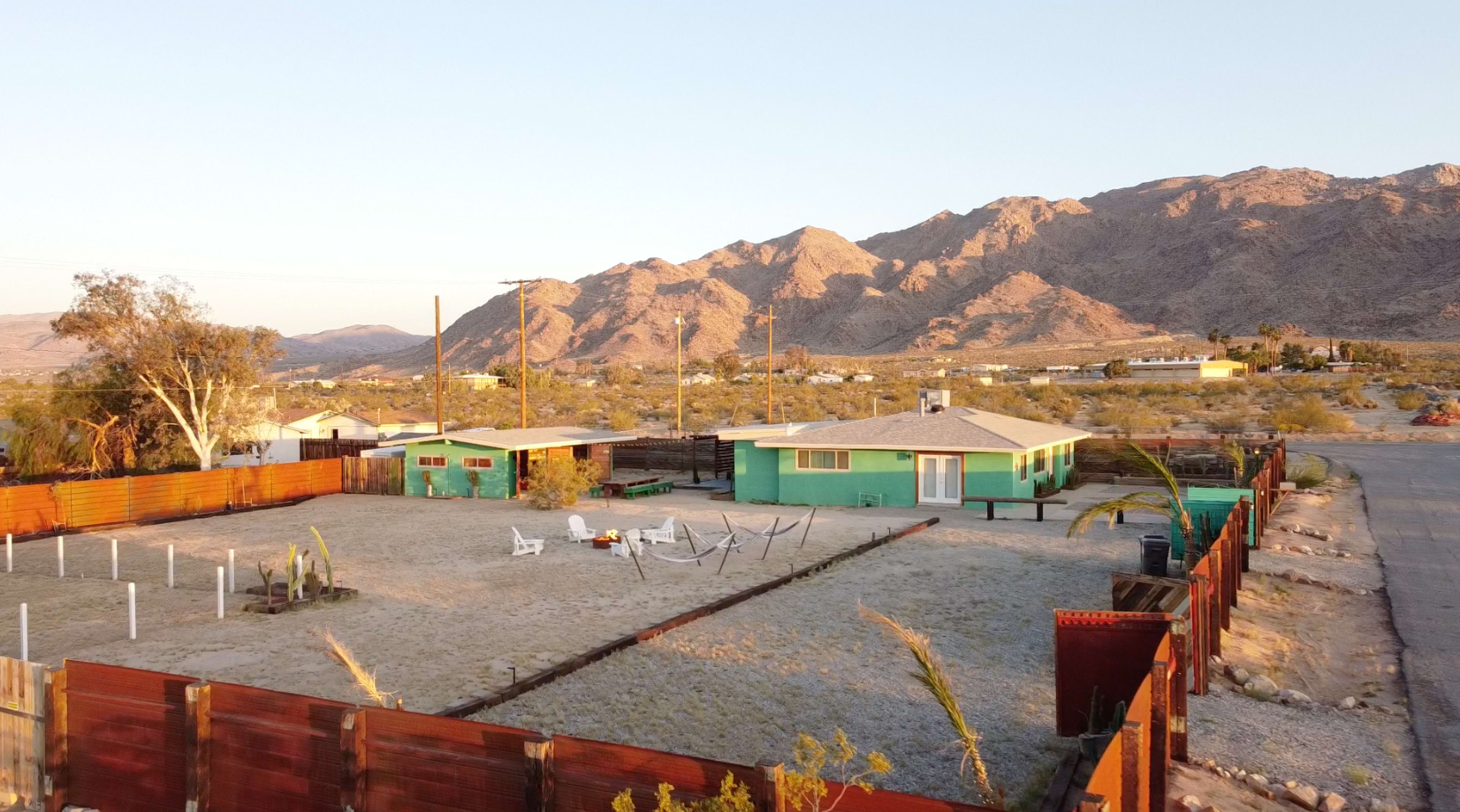 A green house with a sandy yard and white lounge chairs is surrounded by a wooden fence, with mountains in the background and a clear sky.