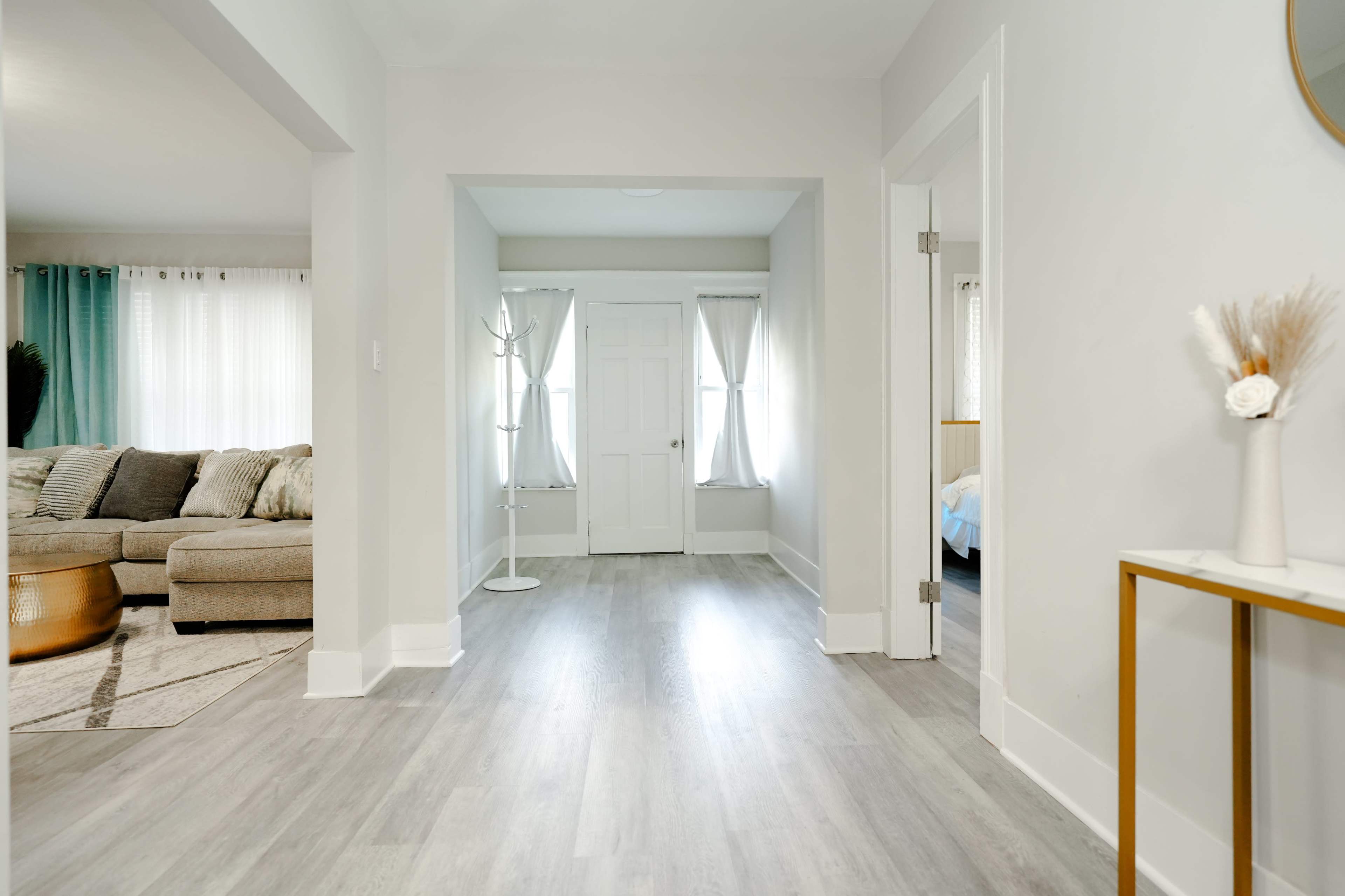 A spacious, well-lit hallway leading to a door and two rooms, with a cozy living area visible on the left.