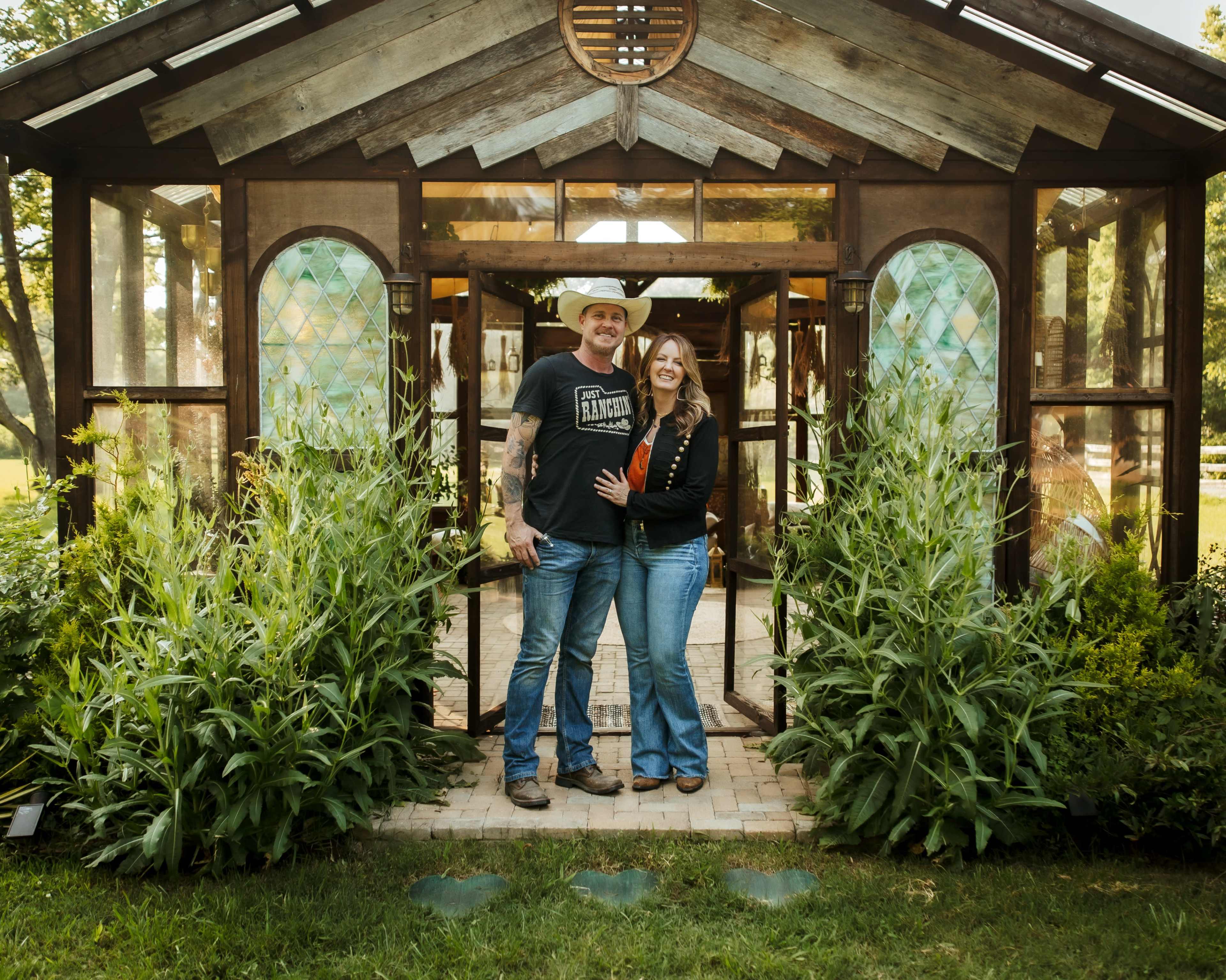 A couple stands together outside a glass greenhouse surrounded by tall greenery.