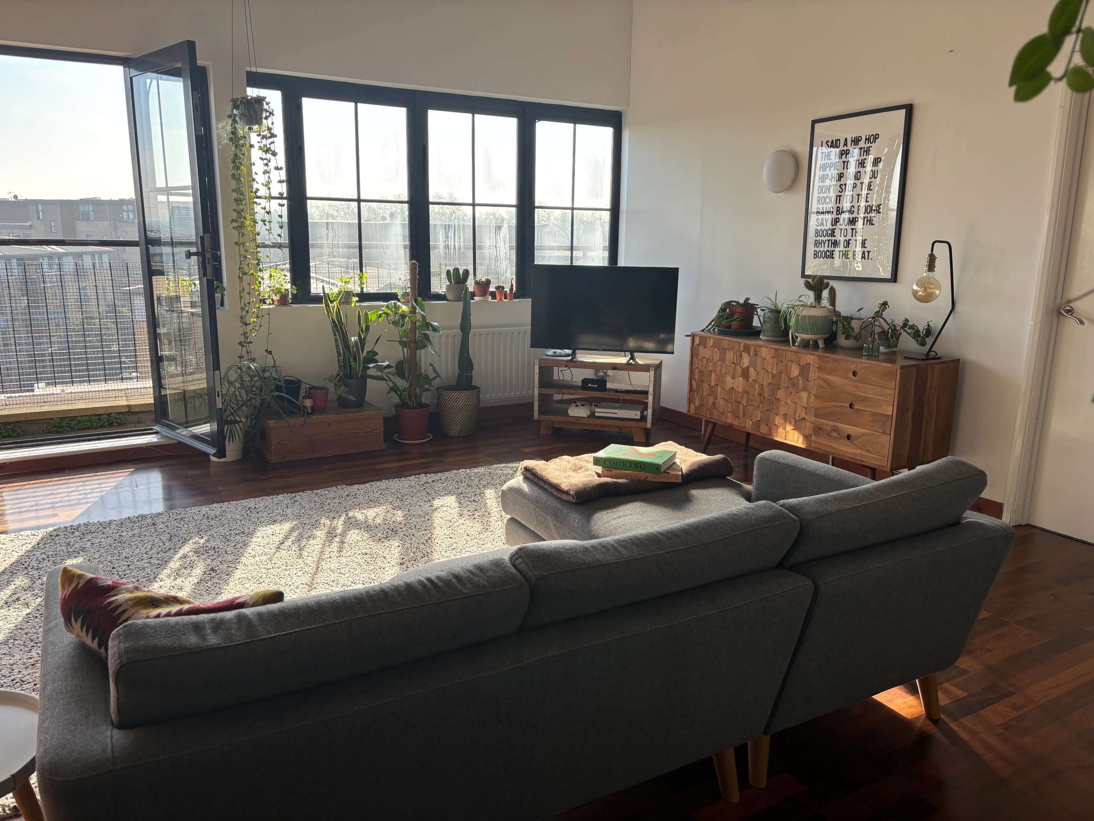 A modern living room features a gray sofa, a wooden cabinet, a television, and large windows overlooking a balcony filled with plants.