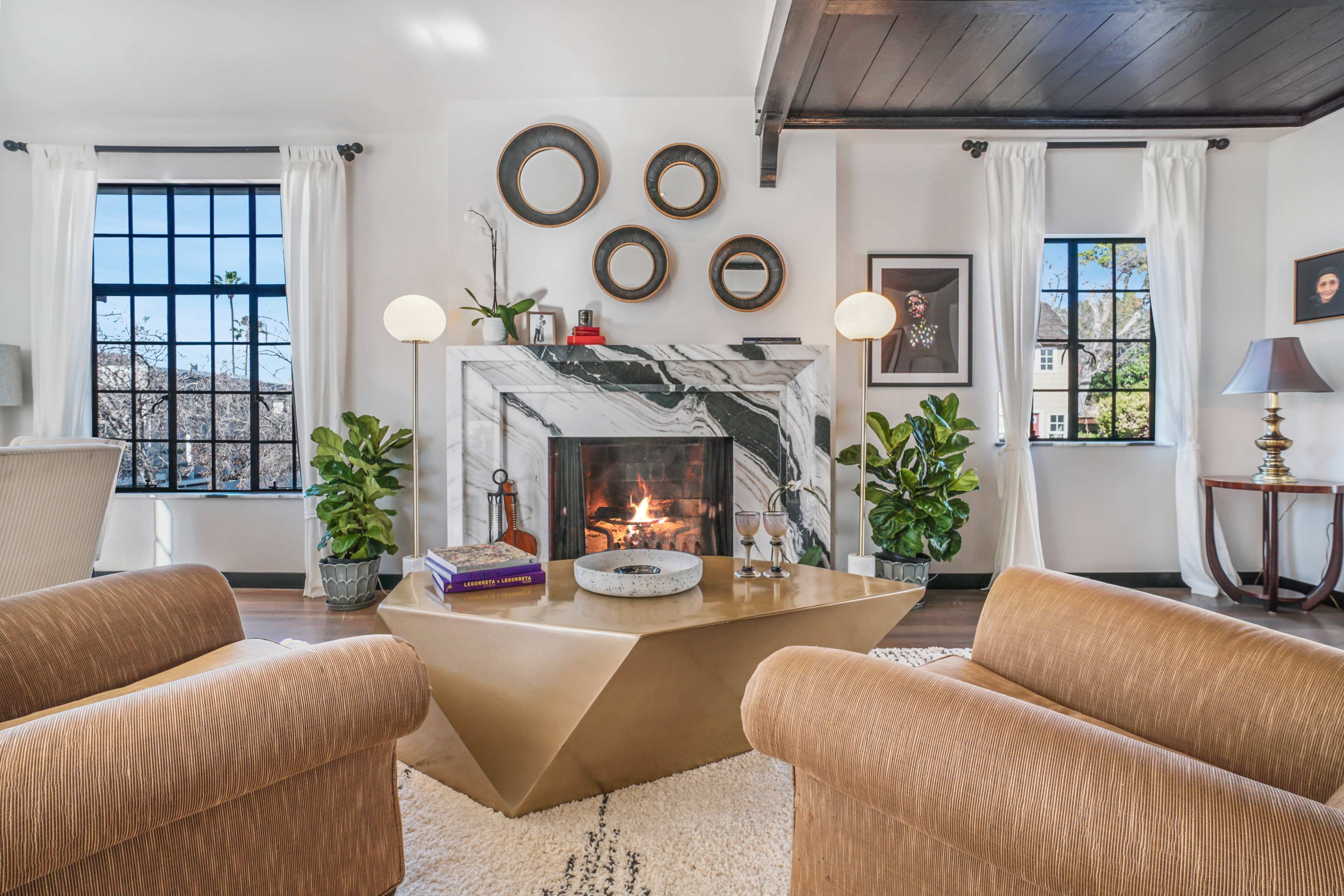 A modern living room features a marble fireplace, gold coffee table, and large windows with plants on either side.