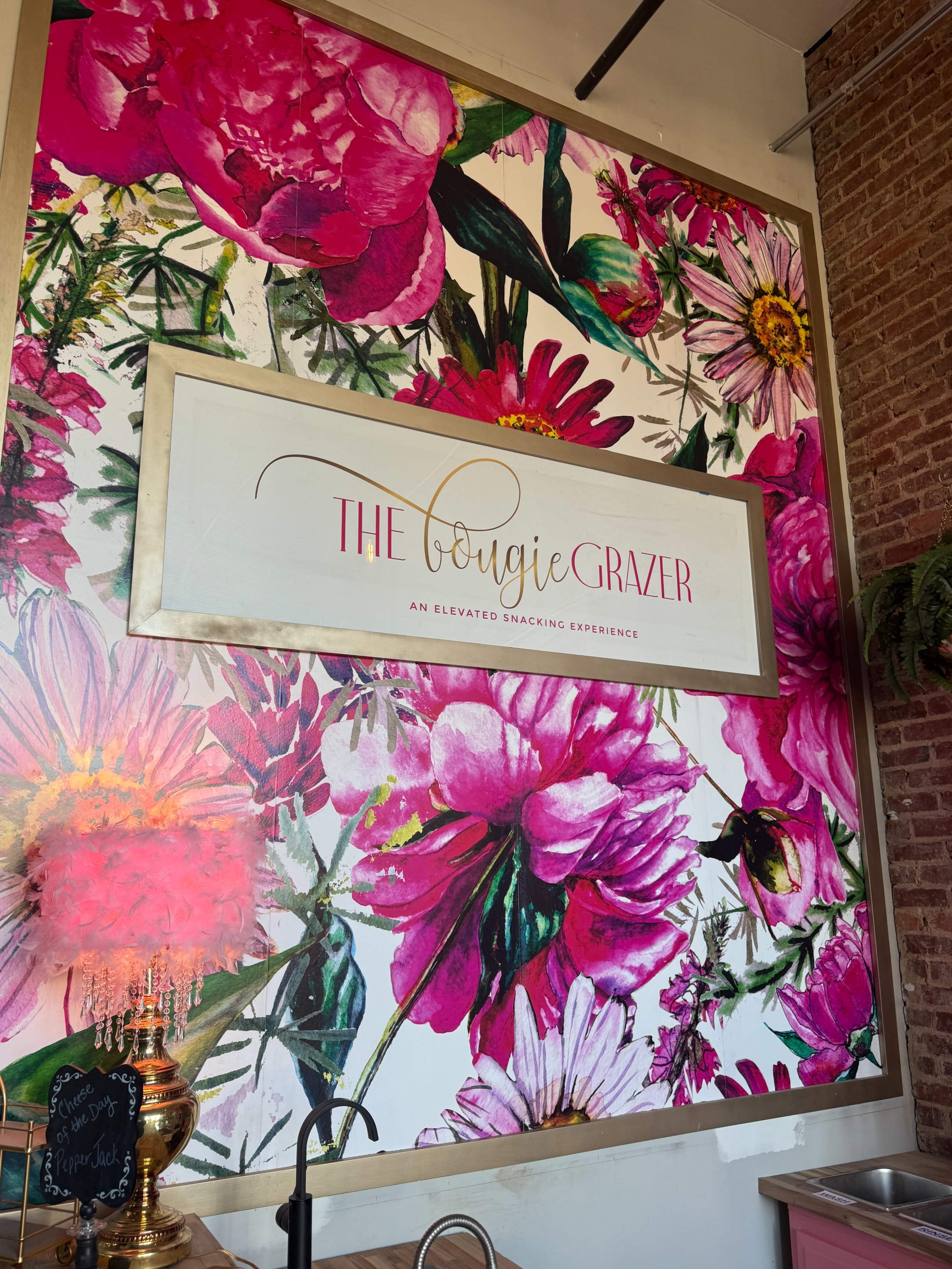 The image features a brightly-colored floral mural with the text "THE Bougie Grazer" prominently displayed in the center, promoting an elevated snacking experience.