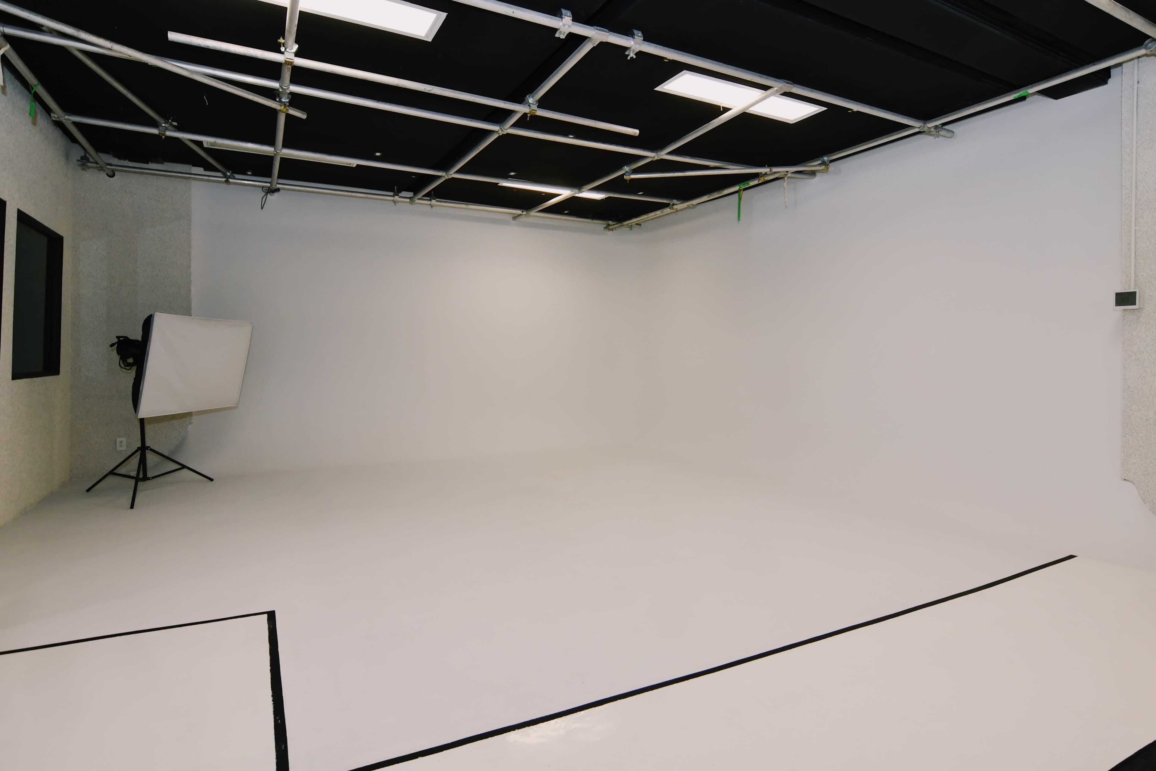 A wide, empty studio space with a white floor and walls, featuring a grid ceiling and minimal furniture.