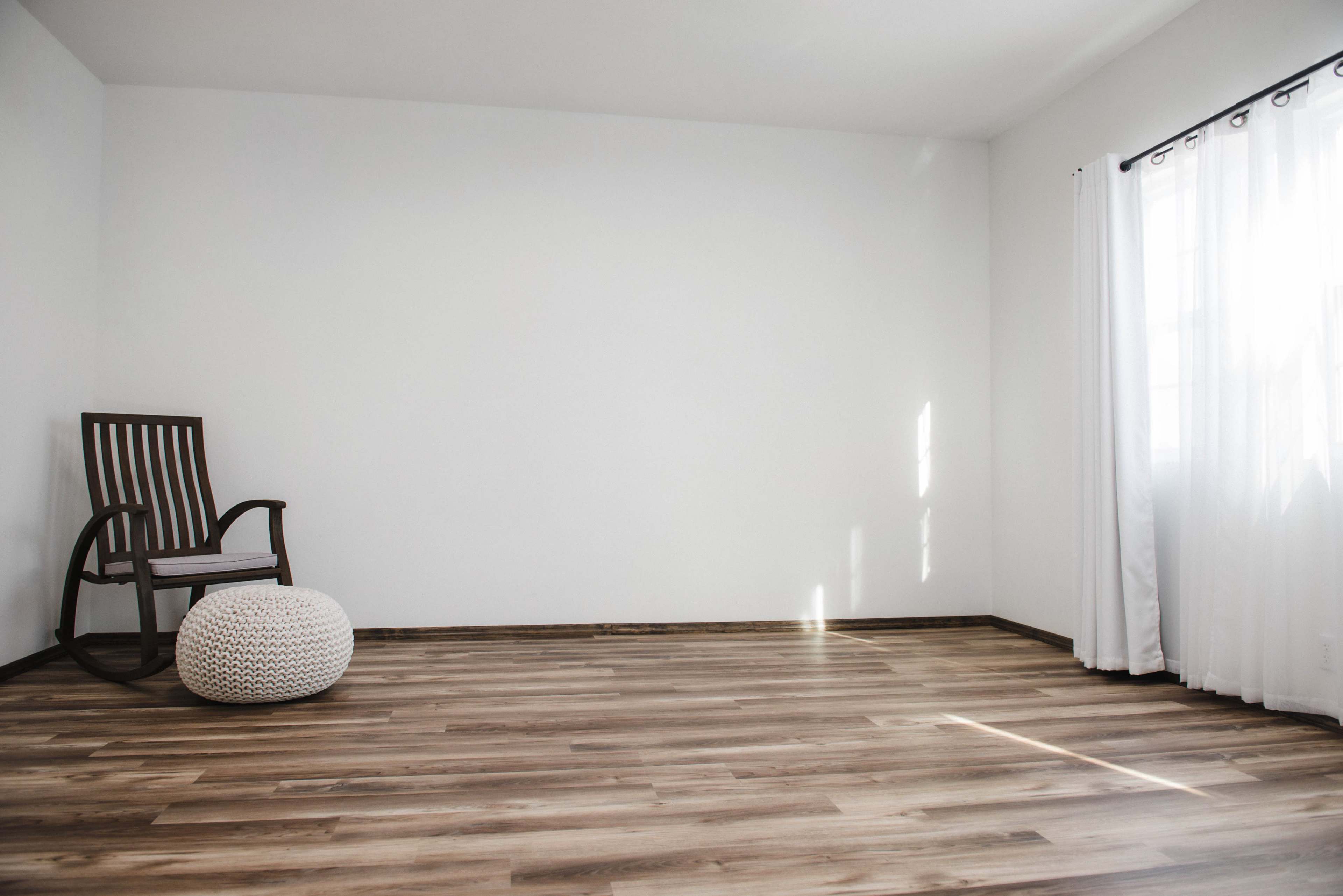 An empty room with light wooden flooring, a rocking chair in one corner, and a white pouf near the chair, accompanied by sheer curtains on the window.