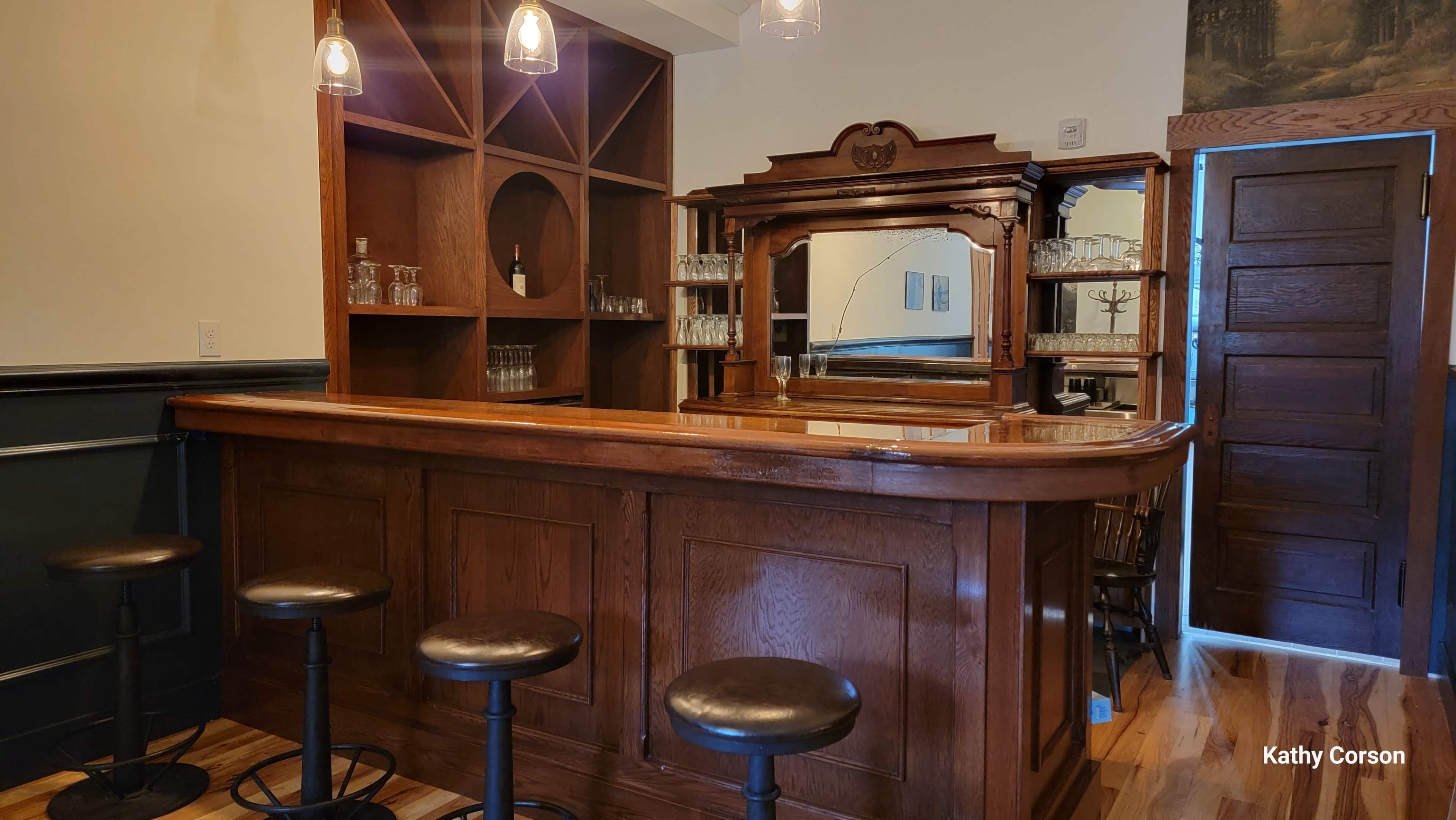 The image shows an elegant wooden bar with a mirror and glassware on shelves, accompanied by several high stools.