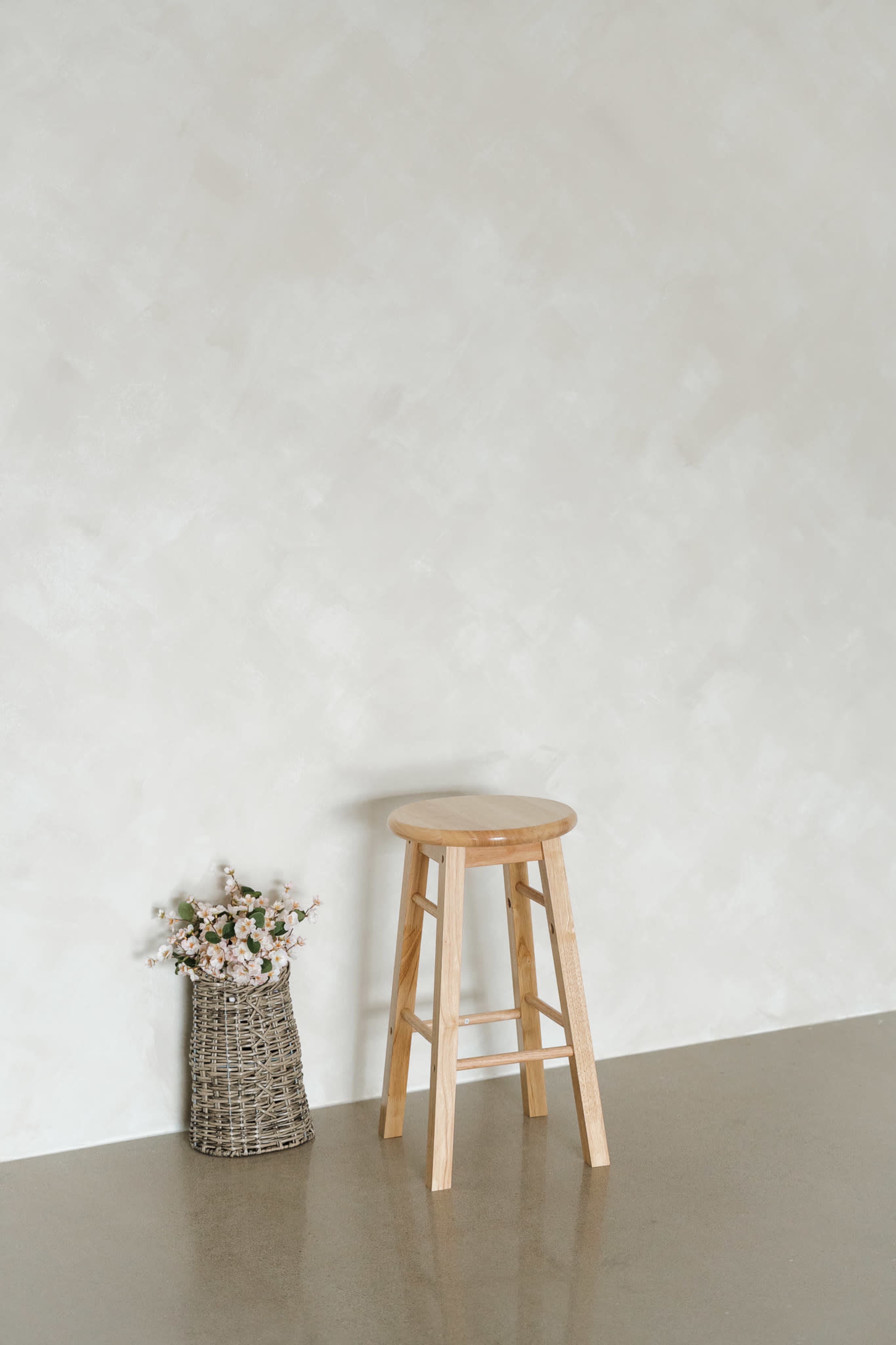 A wooden stool stands next to a woven basket filled with flowers against a light-colored textured wall.