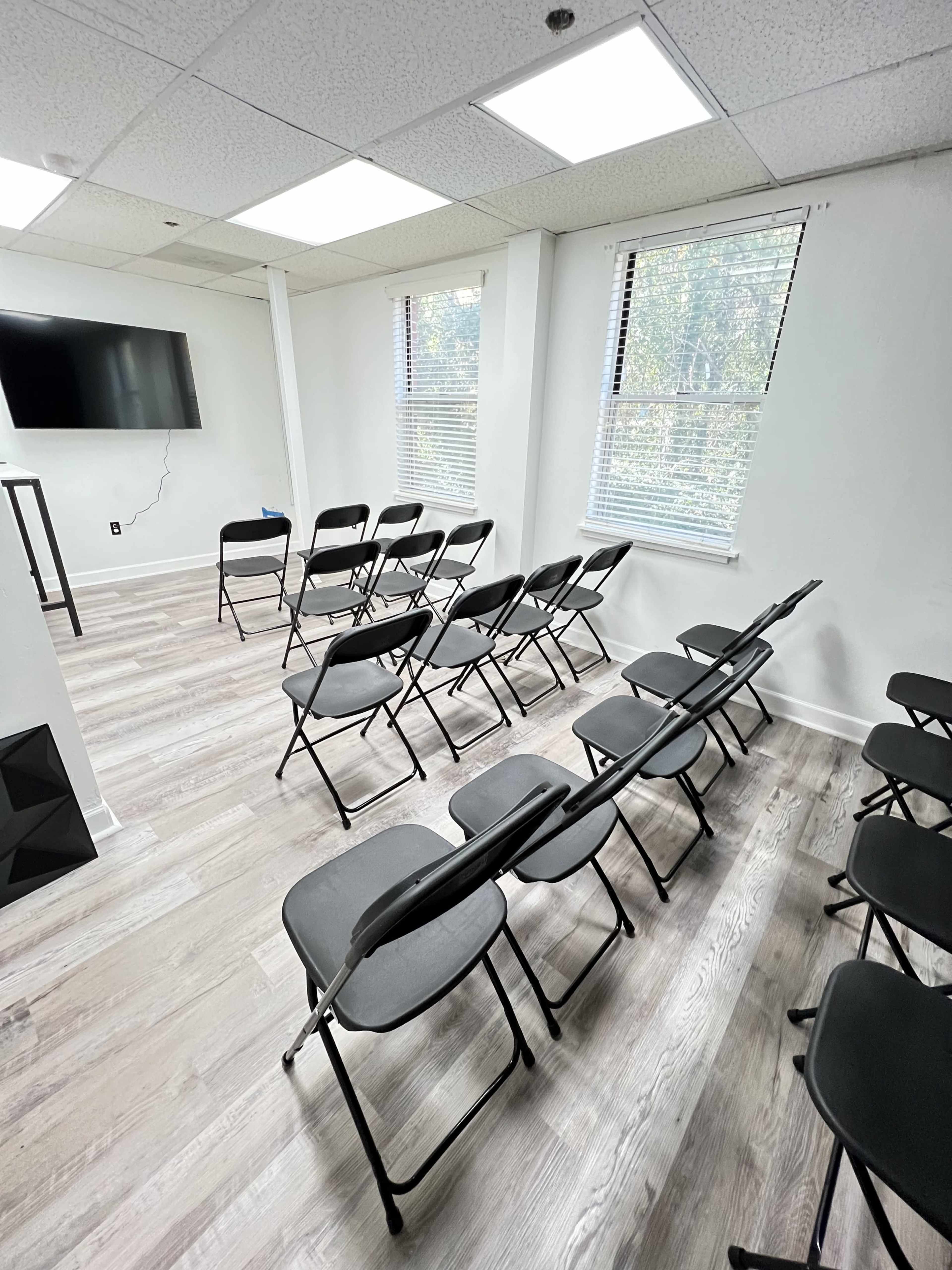 A white-walled room features several black folding chairs arranged in two rows facing a wall-mounted television.