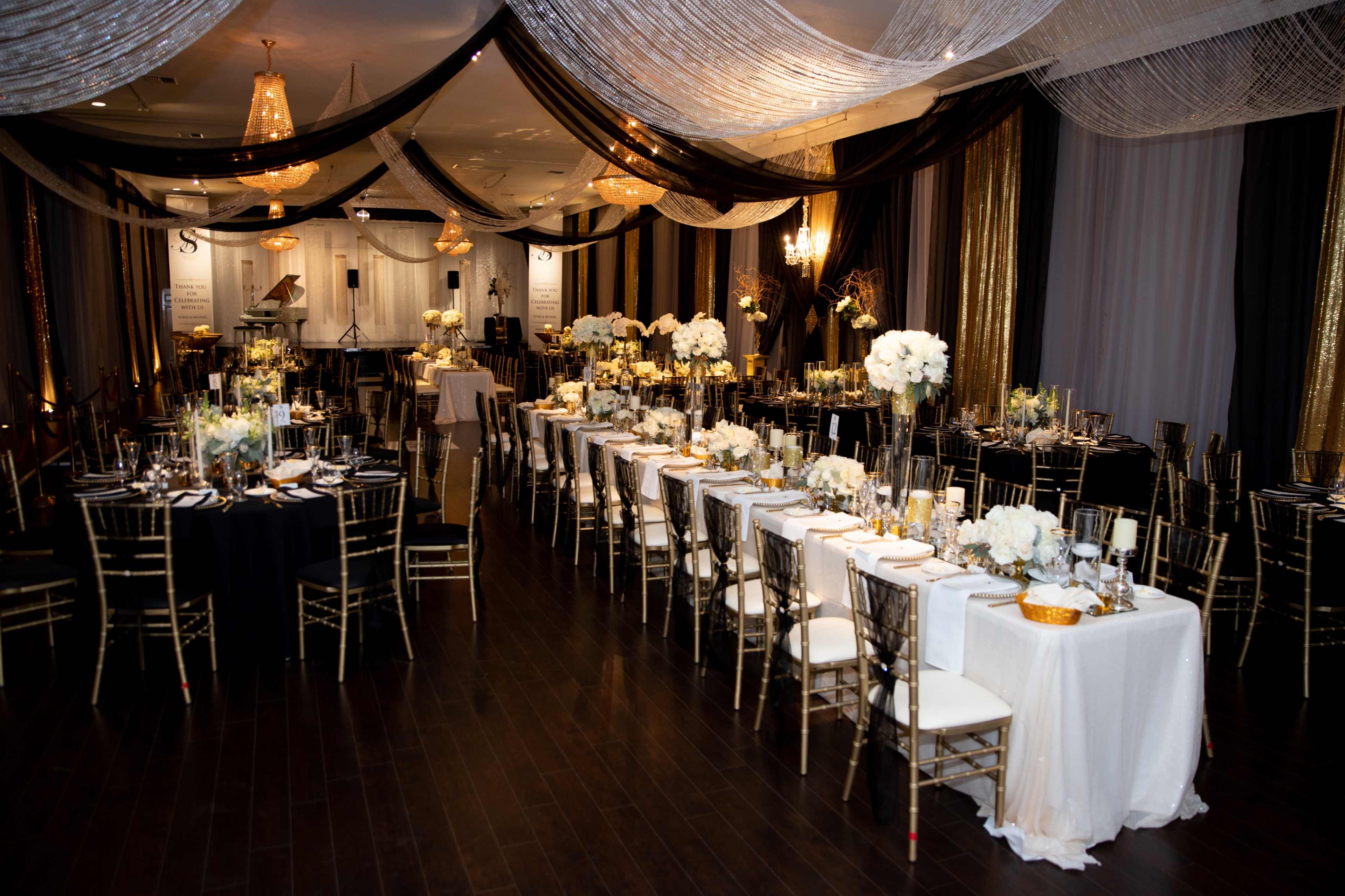 A spacious banquet hall is set up for a formal event, featuring elegantly decorated tables with white tablecloths, floral centerpieces, and gold-accented chairs under draped fabric and hanging chandeliers.