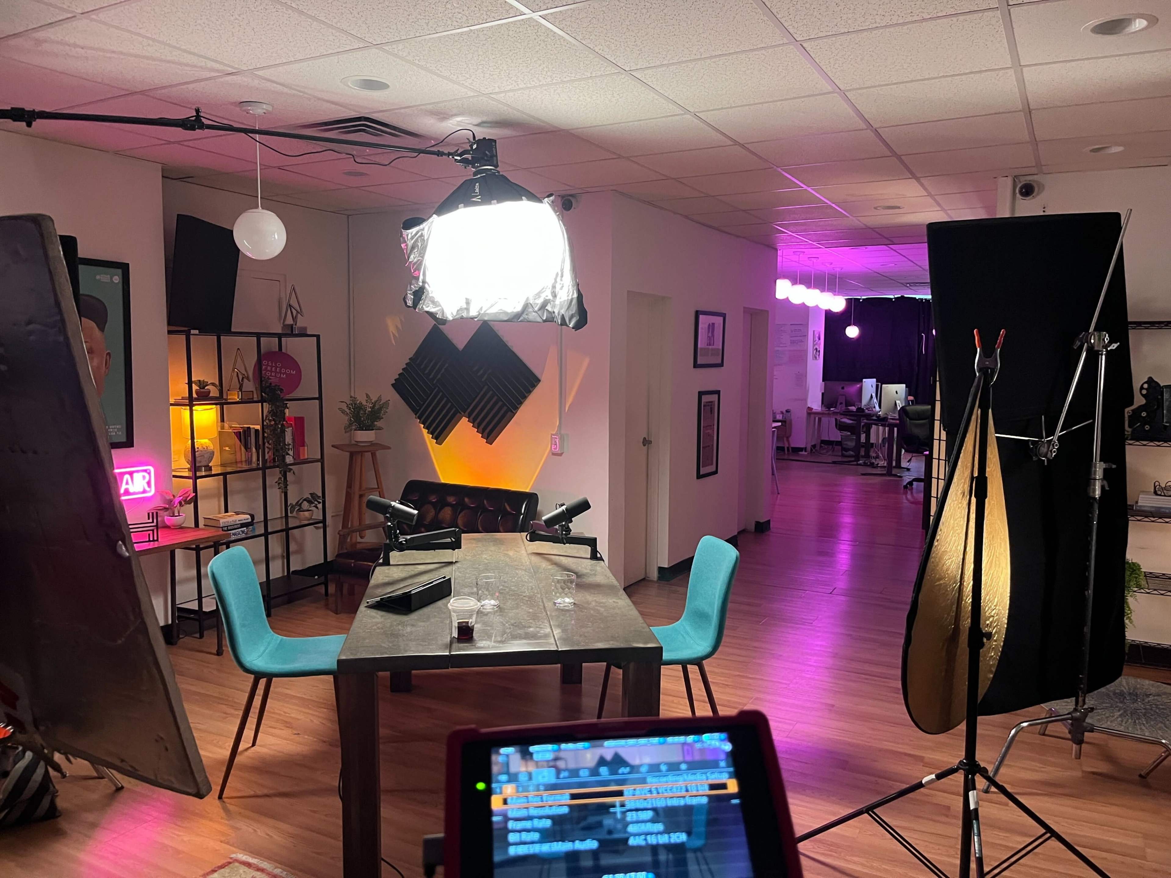The image shows a well-lit studio space featuring a table with chairs, a backdrop setup, and a bright light source in an office-like environment.
