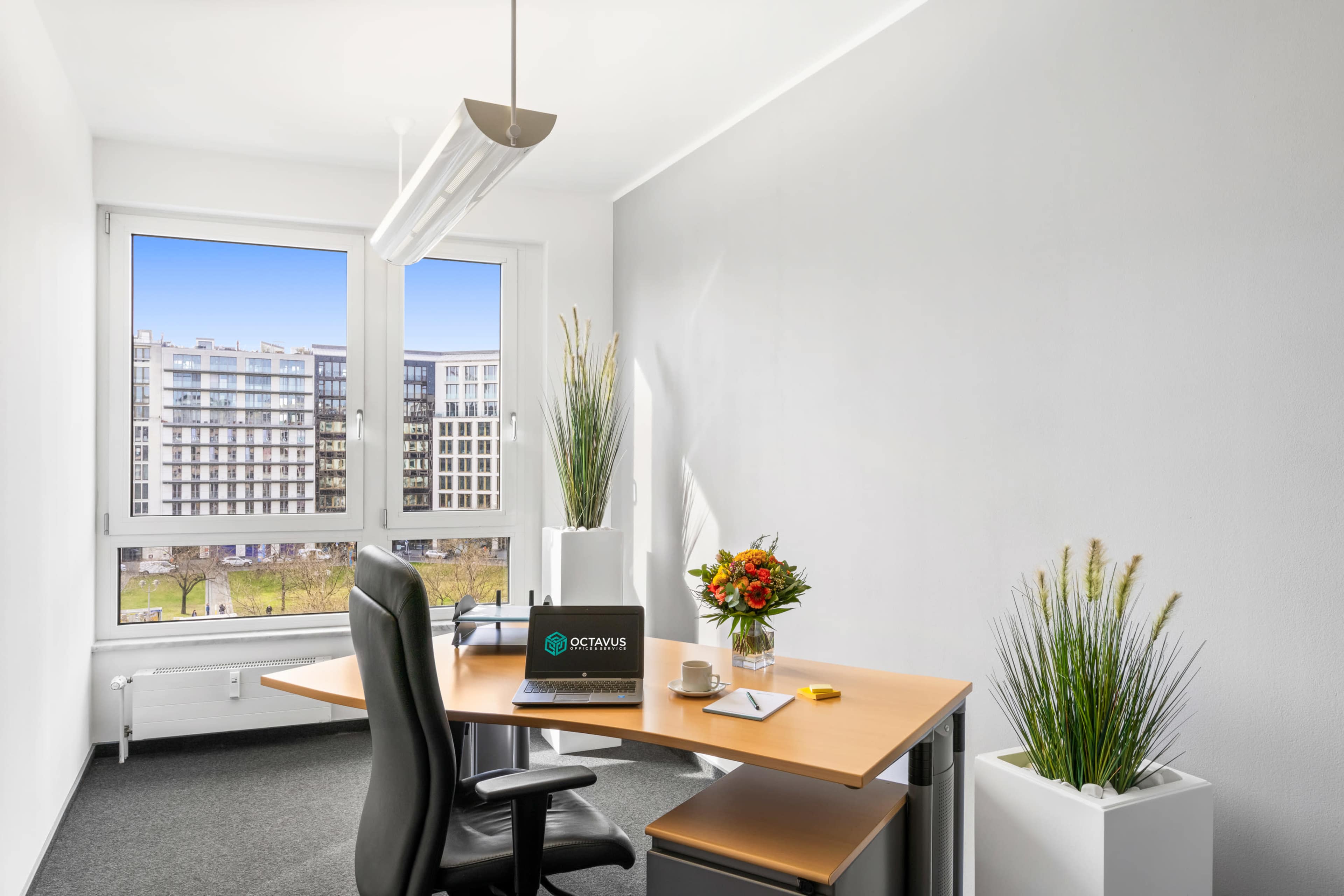 A modern office space features a desk with a laptop, a small decorative flower arrangement, and large windows overlooking a landscaped area.