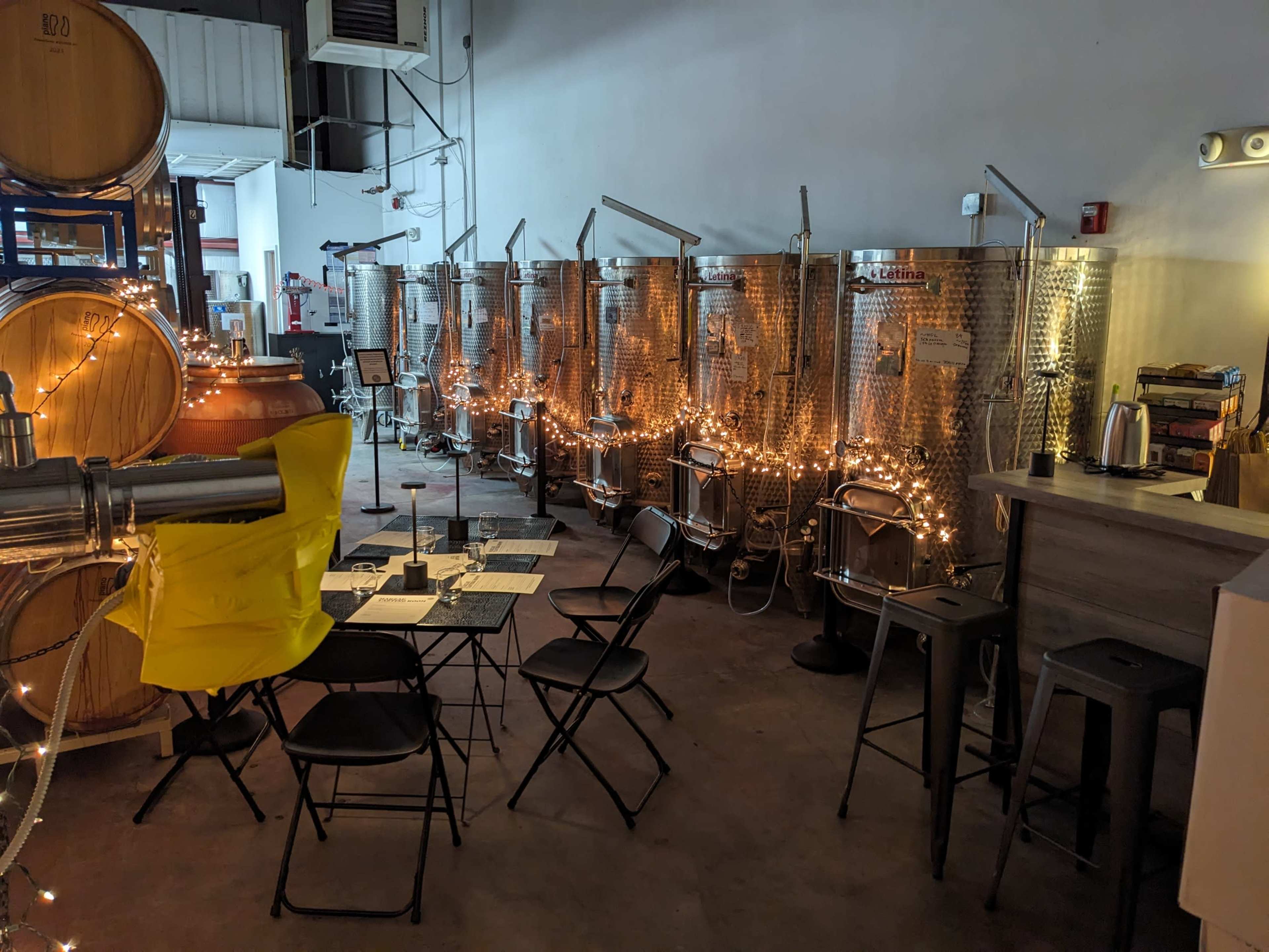 A dimly lit room with stainless steel fermentation tanks lined against a wall, string lights illuminating the space, and a seating area with tables and chairs.