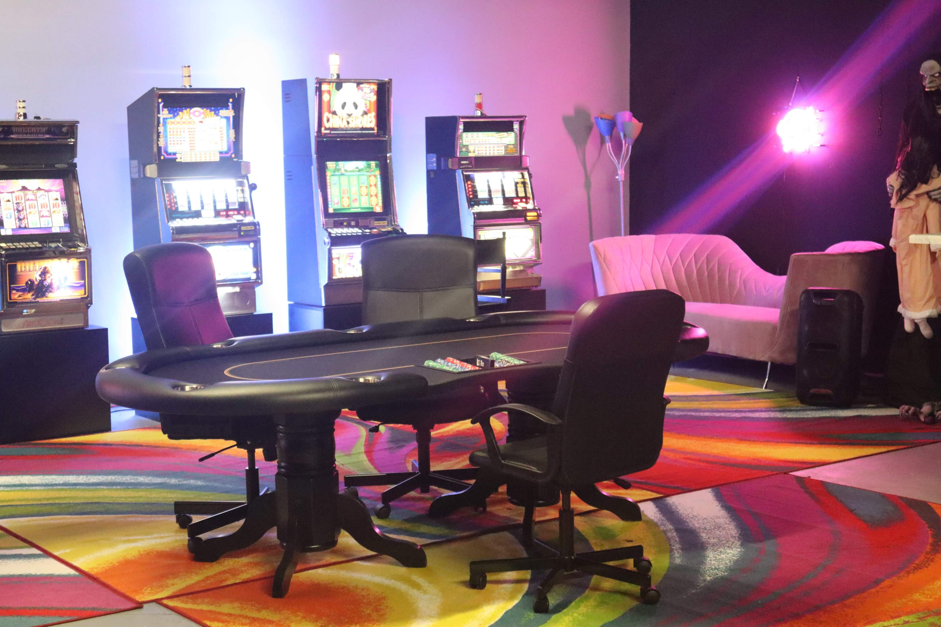 A poker table surrounded by chairs is set in a colorful room with slot machines in the background and a sofa on one side.