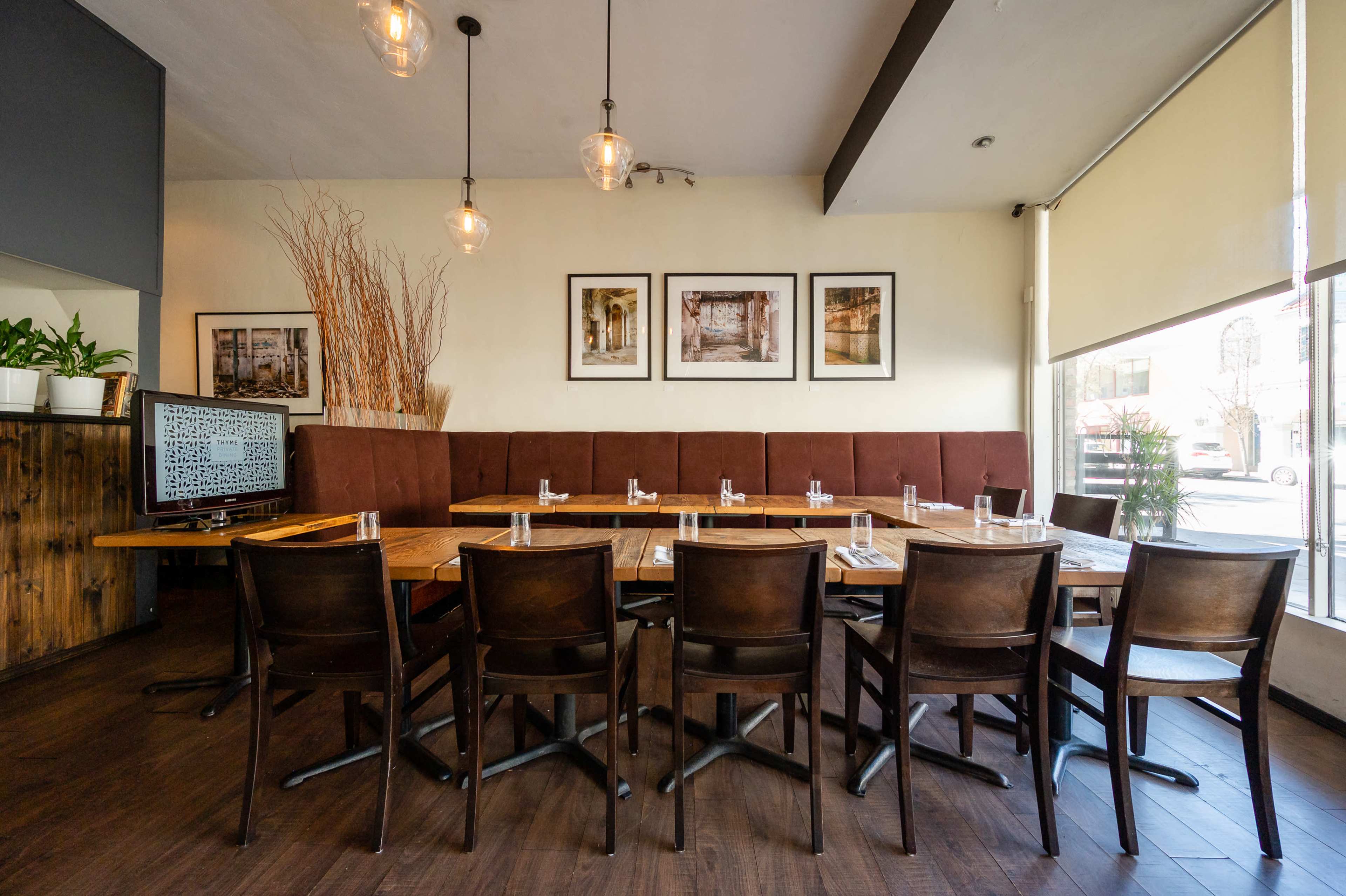 A modern restaurant interior with a long table set for dining, surrounded by dark wooden chairs and decor featuring framed pictures on the wall.