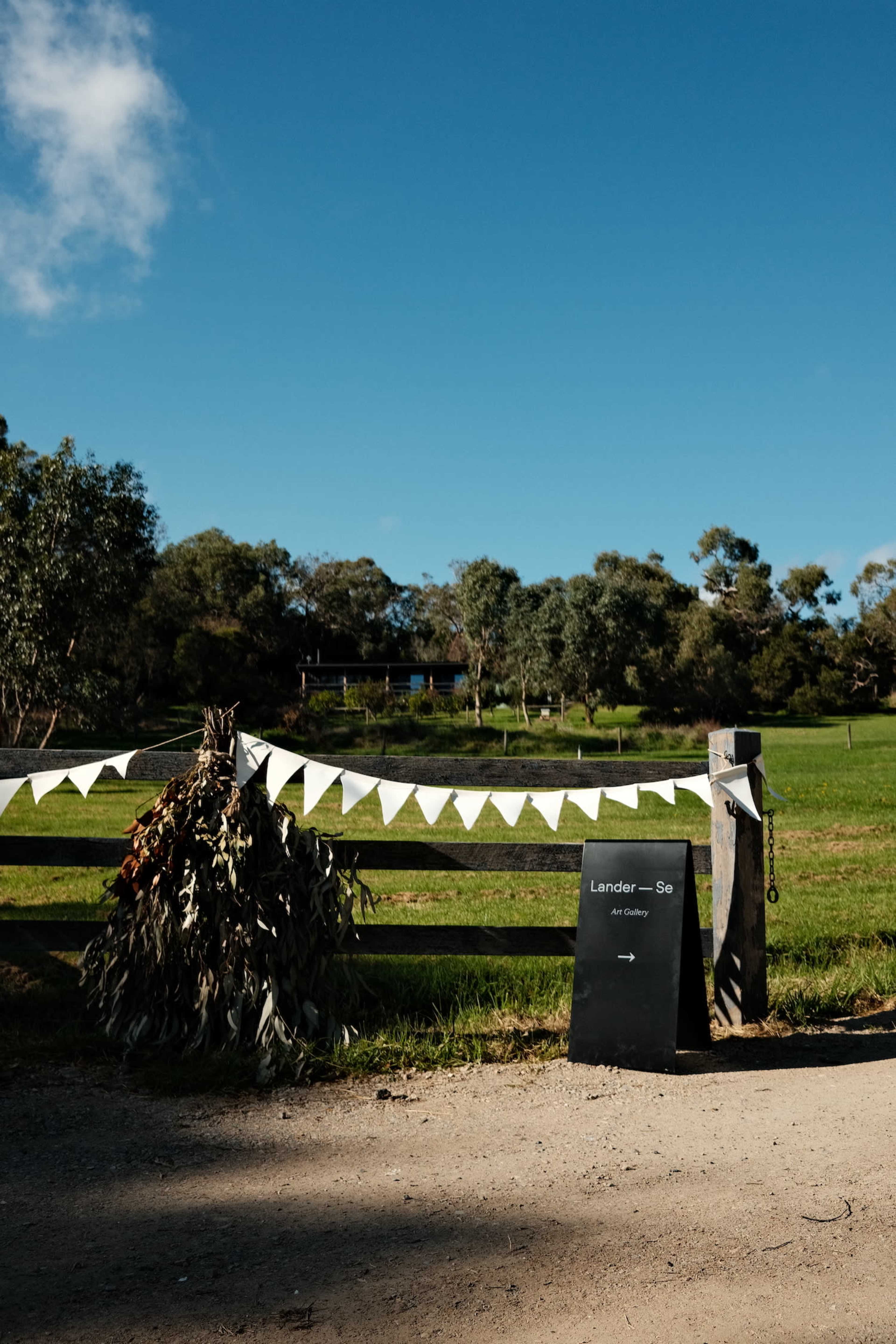 A wooden fence with bunting and a sign marks the entrance to a rural property surrounded by greenery.