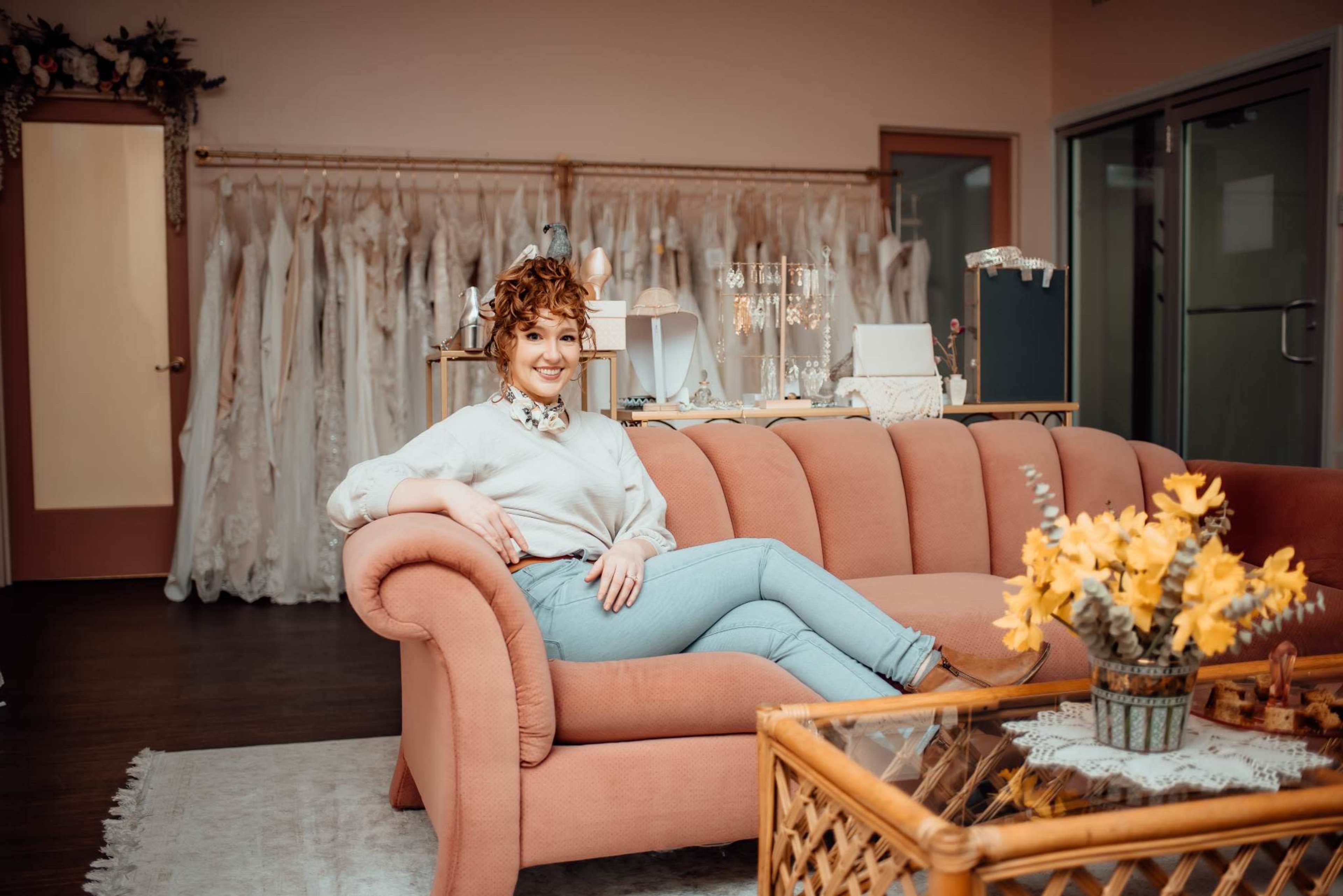 A woman with curly hair sits on a pink couch in a bridal boutique surrounded by wedding dresses hanging in the background.