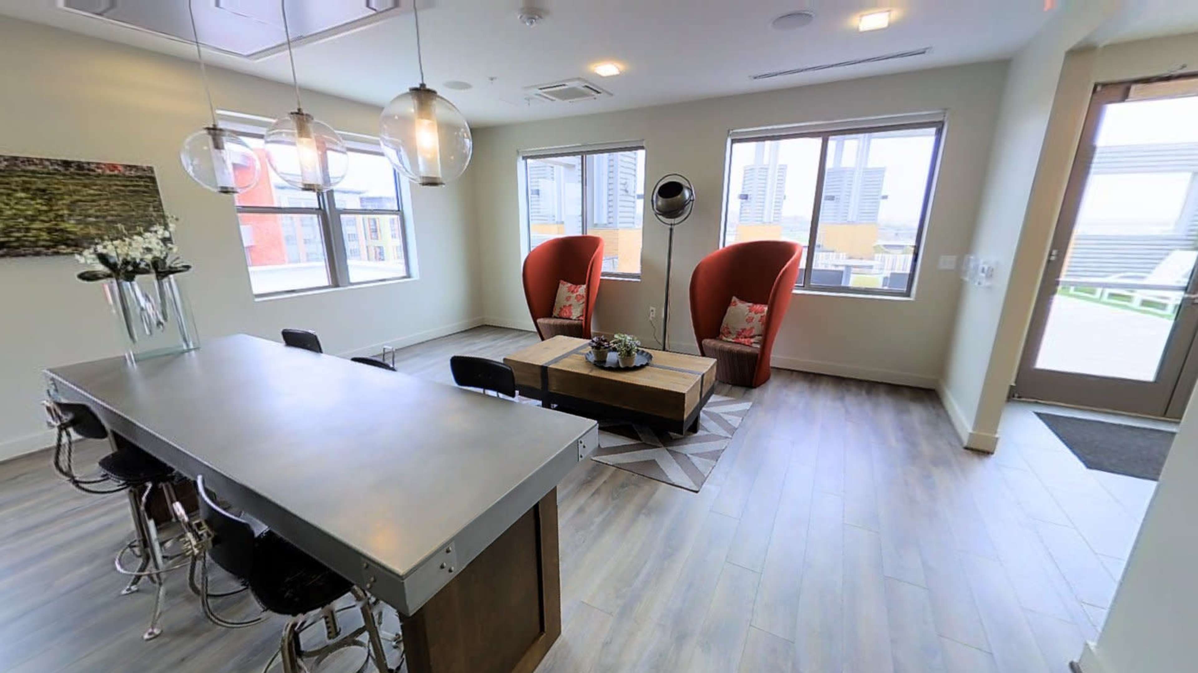 A modern room with two large red chairs, a wooden coffee table, a dining area with six black chairs, and large windows overlooking a cityscape.
