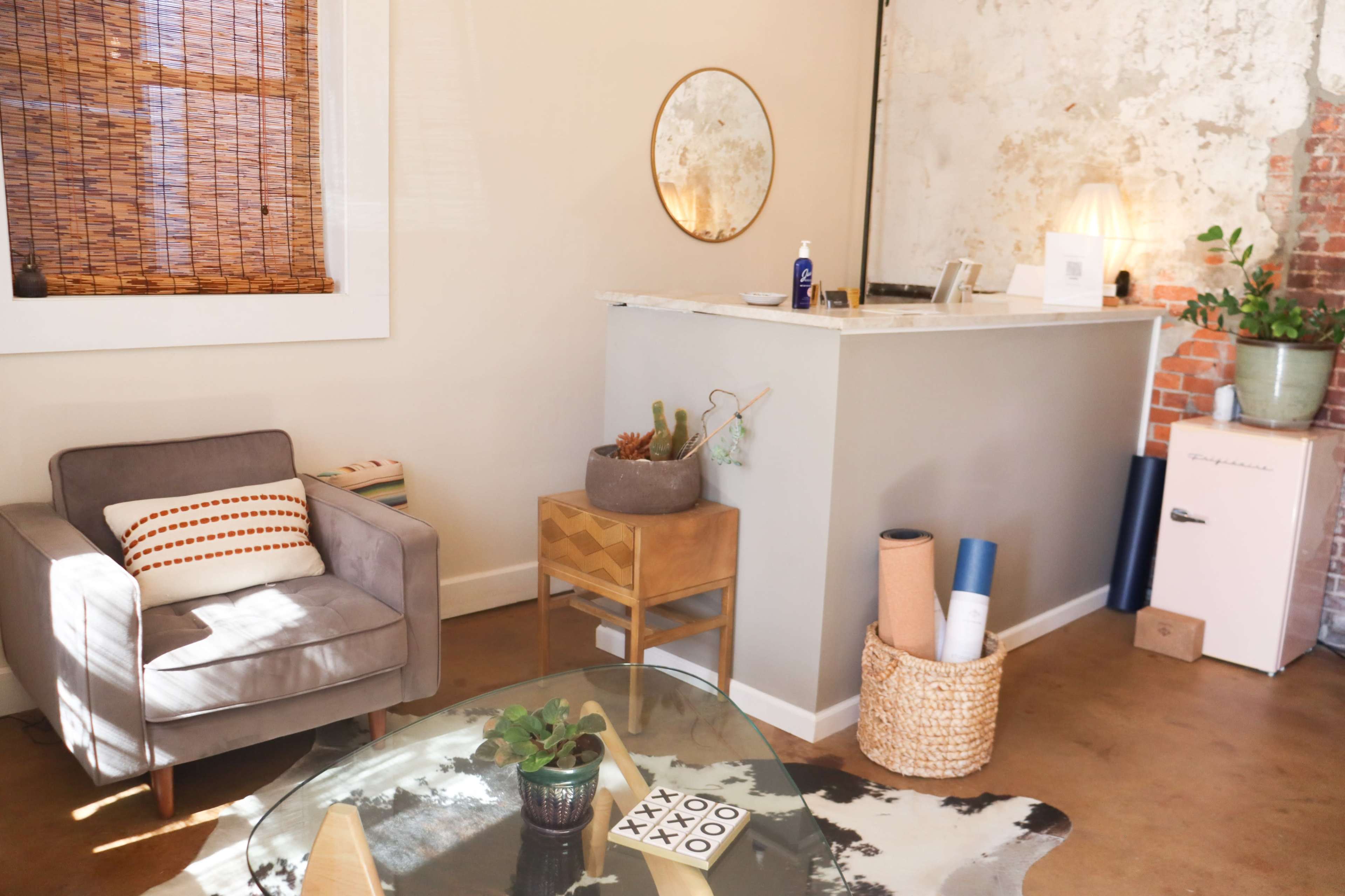 A cozy interior with a gray armchair, a small wooden table, a minimalist reception area with a round mirror, and decorative plants.