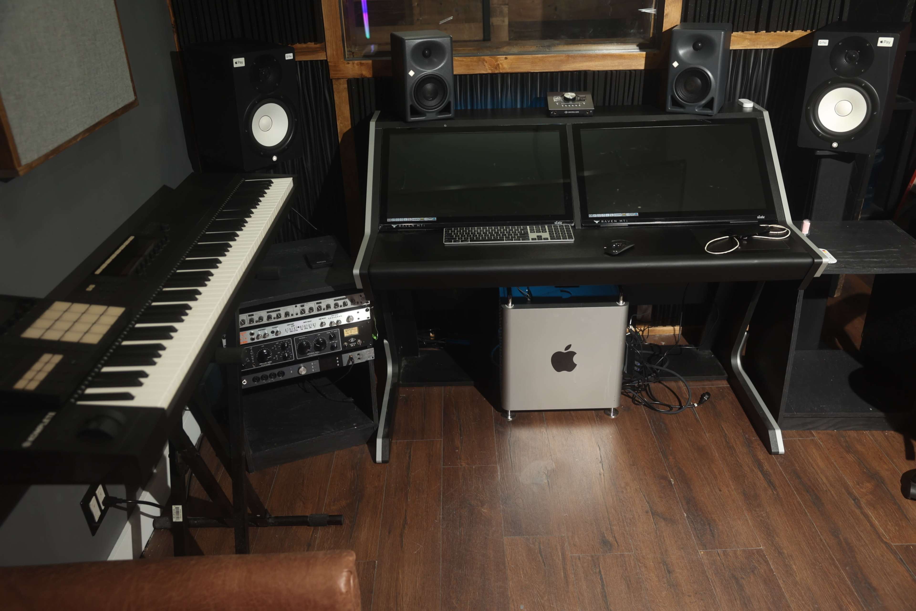 A music production workspace with a keyboard, dual computer monitors, speakers, and various audio equipment on wooden flooring.