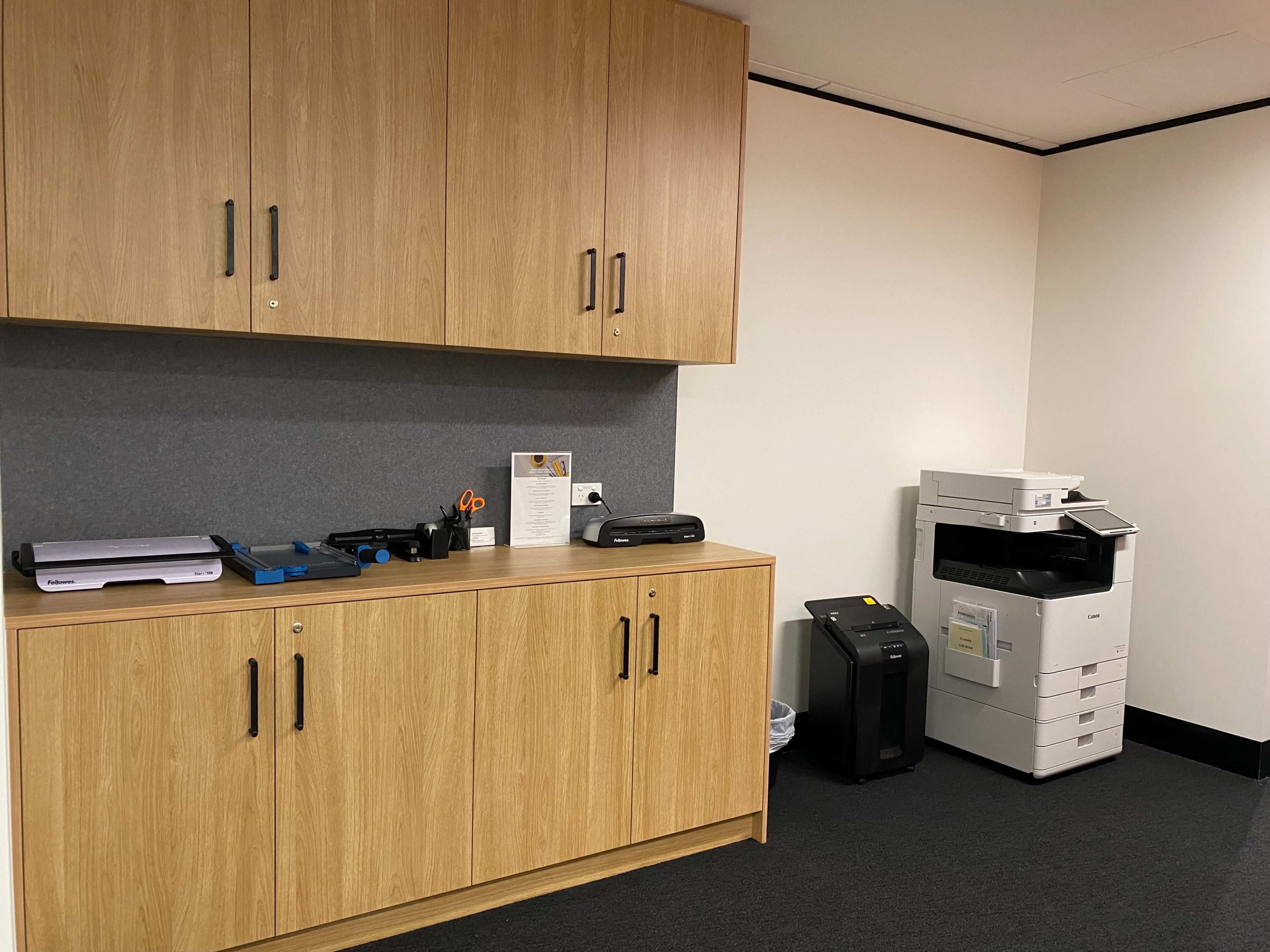 The image shows a small office space featuring a wooden cabinet with various office supplies, a printer, and a multi-function copier in the corner.
