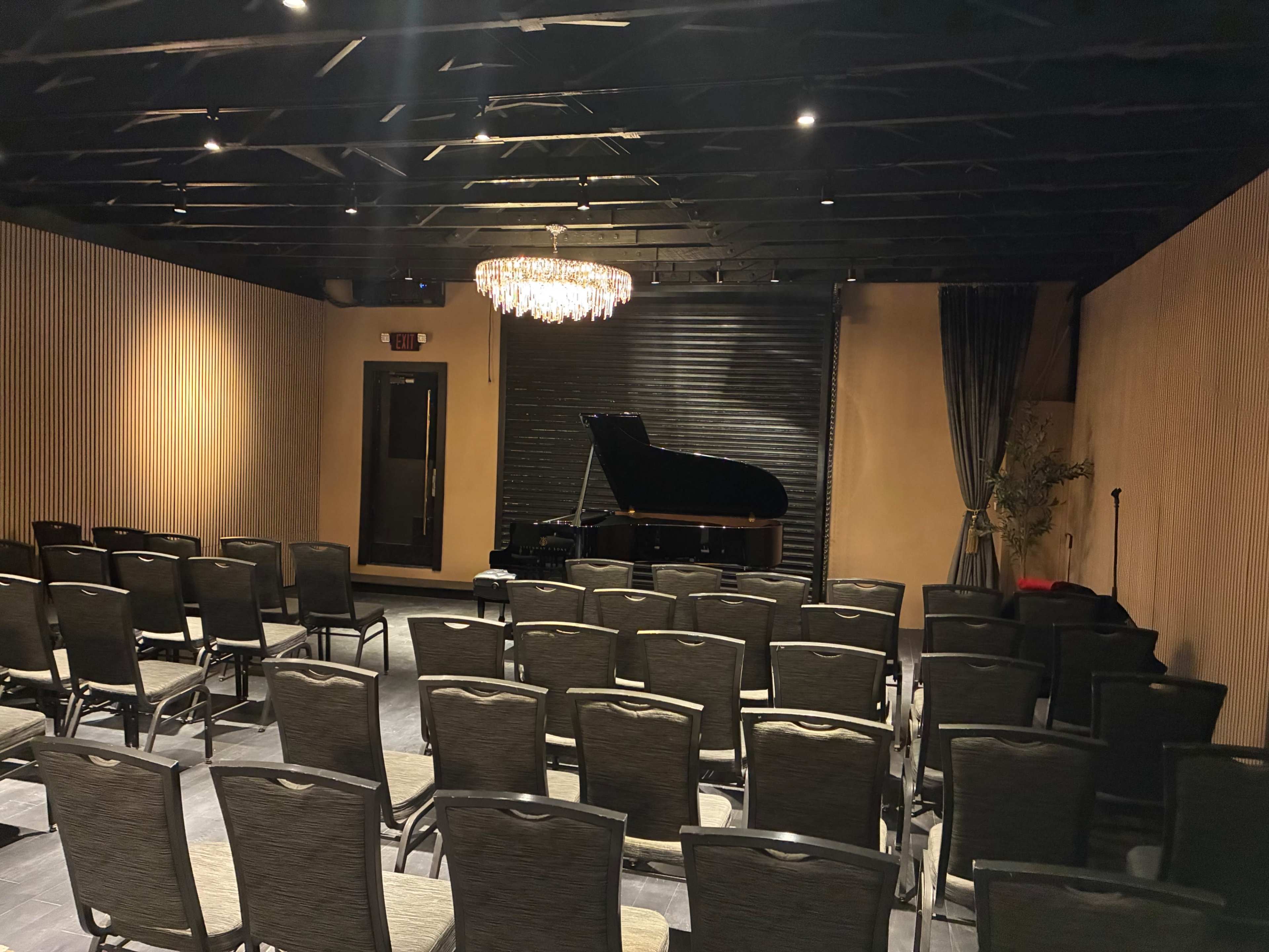 The image shows a dimly lit performance venue with rows of chairs facing a grand piano under a chandelier.