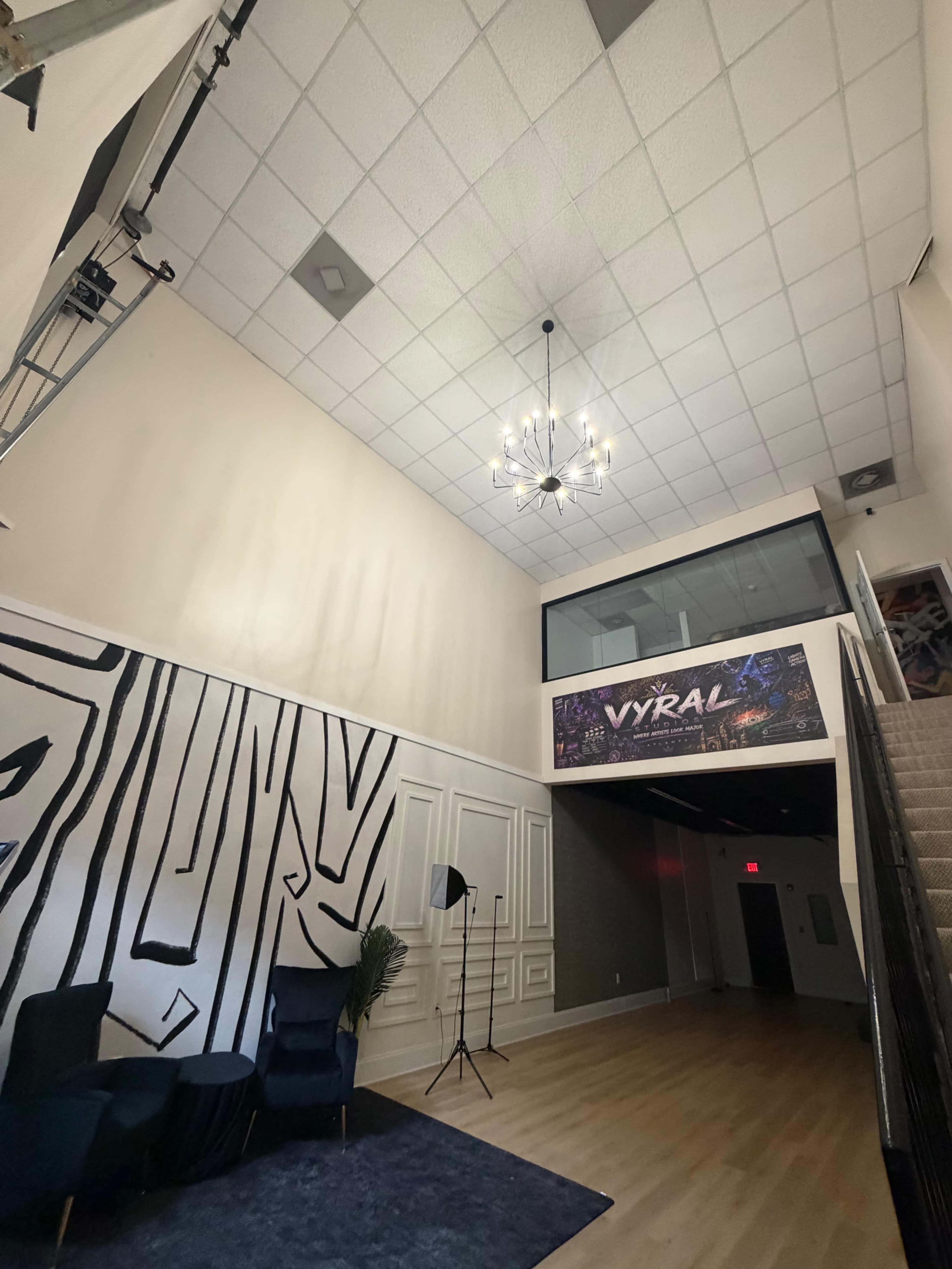 The image shows a spacious lobby with a high ceiling, a chandelier, and a staircase leading to an upper area, featuring a large wall decoration with the word "VYRAL."