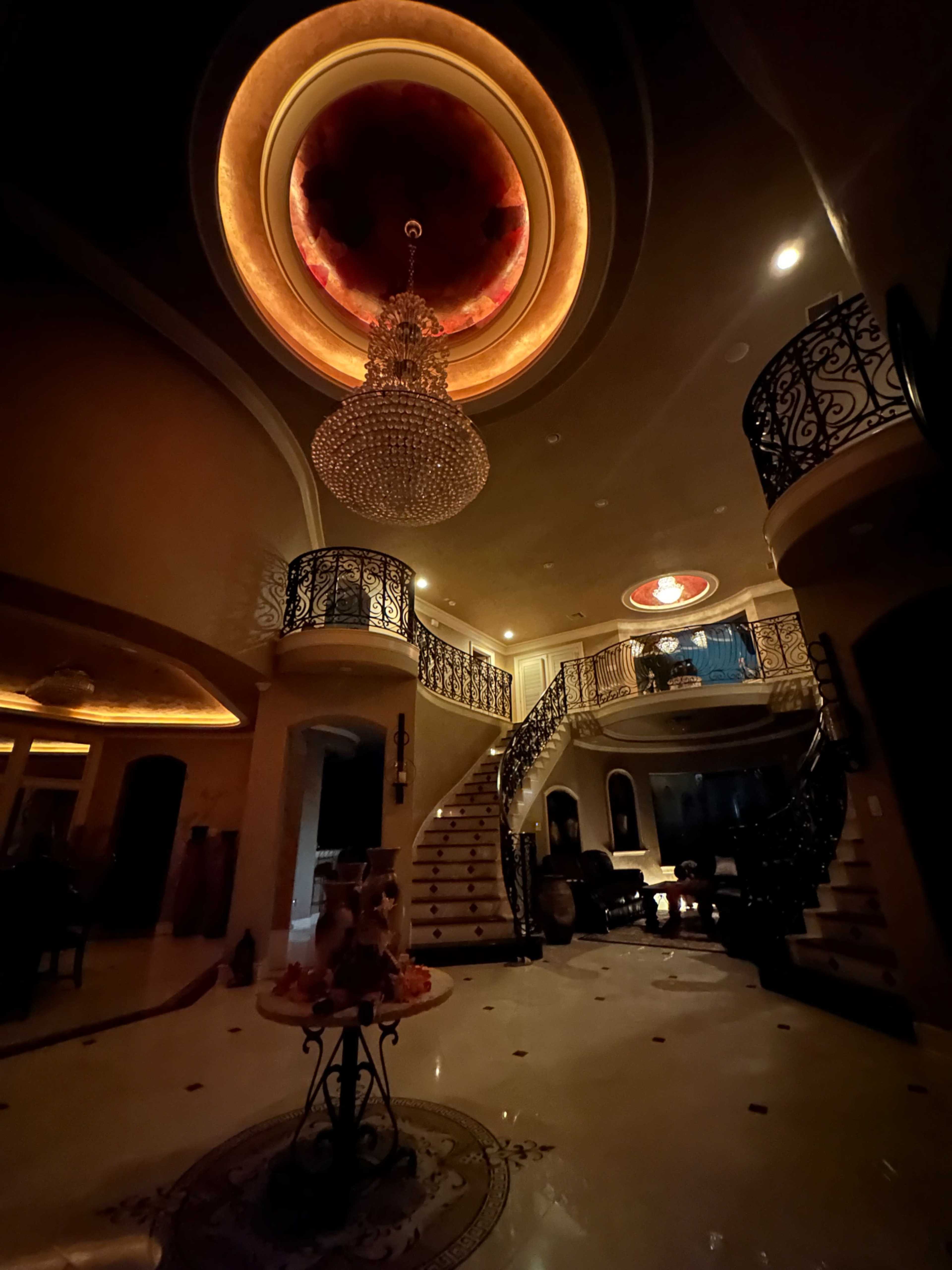 The image shows a luxurious entryway of a home featuring a grand staircase, an ornate chandelier, and decorative ceiling lighting.