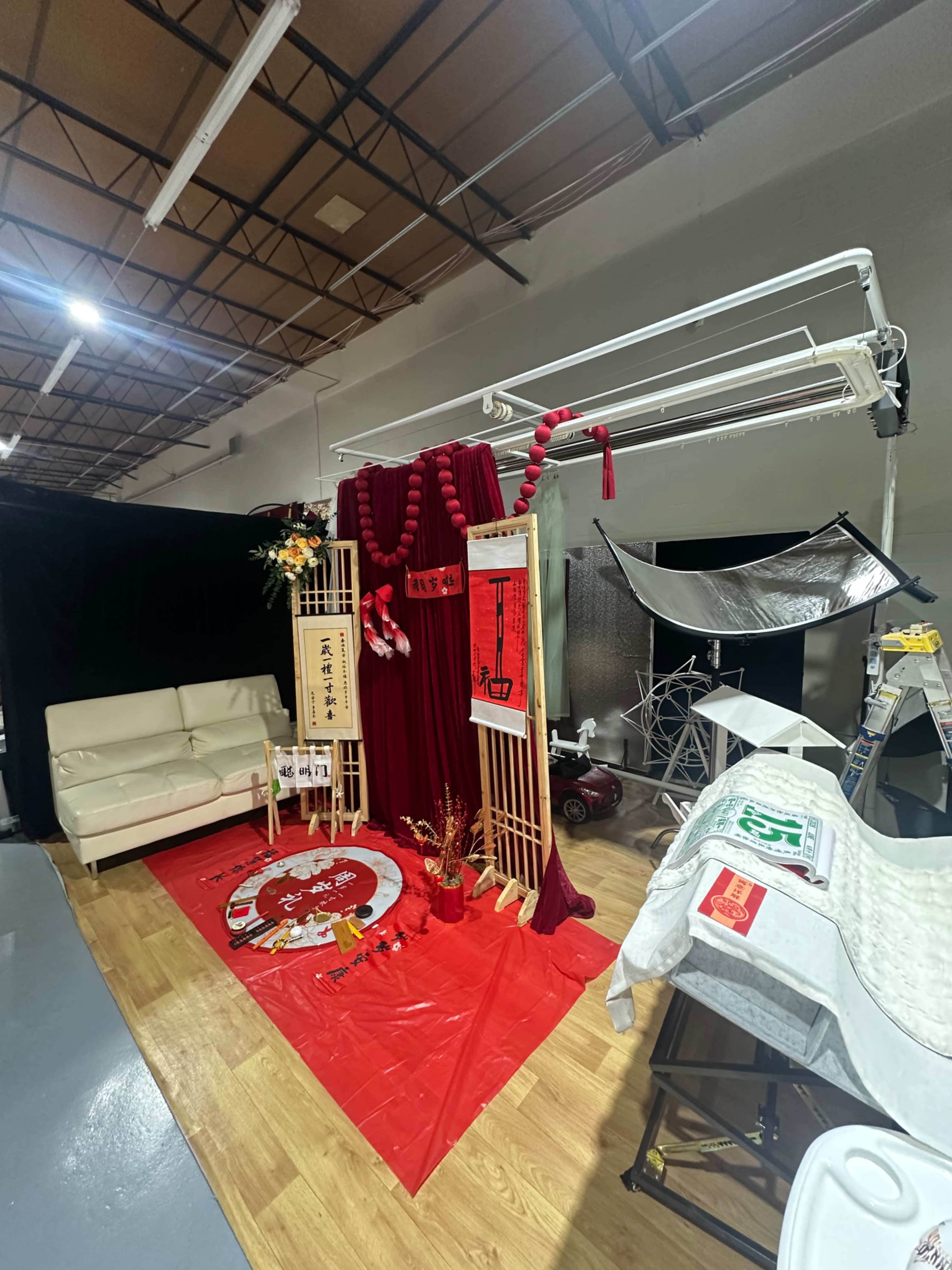 The image shows a decorated studio space featuring red fabric drapes, a white couch, and various props arranged on a wooden floor.