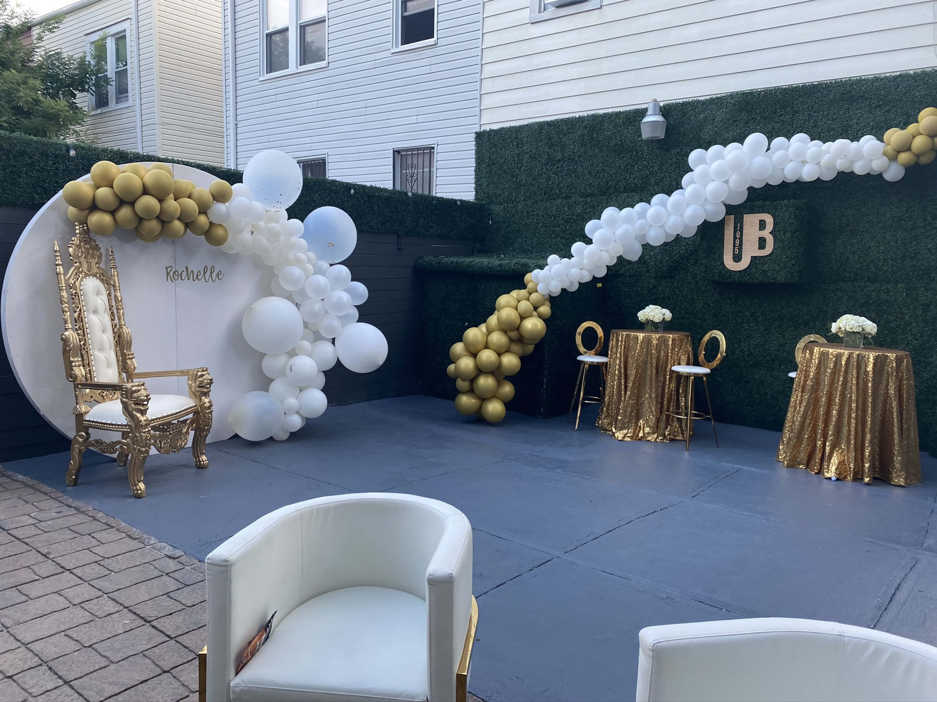 A decorated outdoor space featuring a gold and white balloon arch, a circular backdrop with the name "Michelle," a golden throne chair, and two tables with gold sequined tablecloths.